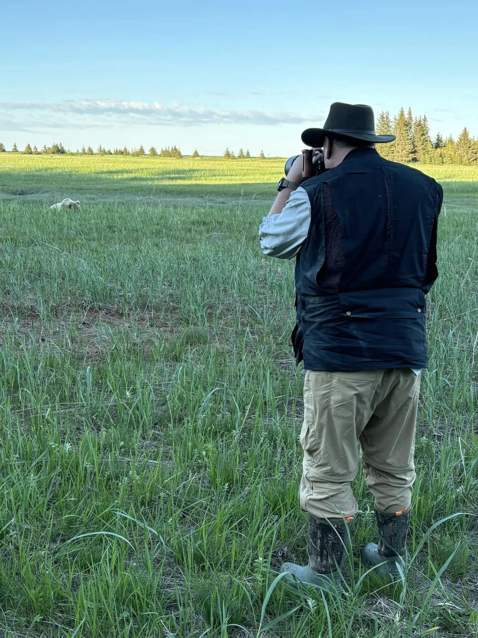 A person in outdoor gear, including a wide-brimmed hat, vest, khaki pants, and rubber boots, is taking a photograph of a sheep in a grassy field with trees and a blue sky in the background.
