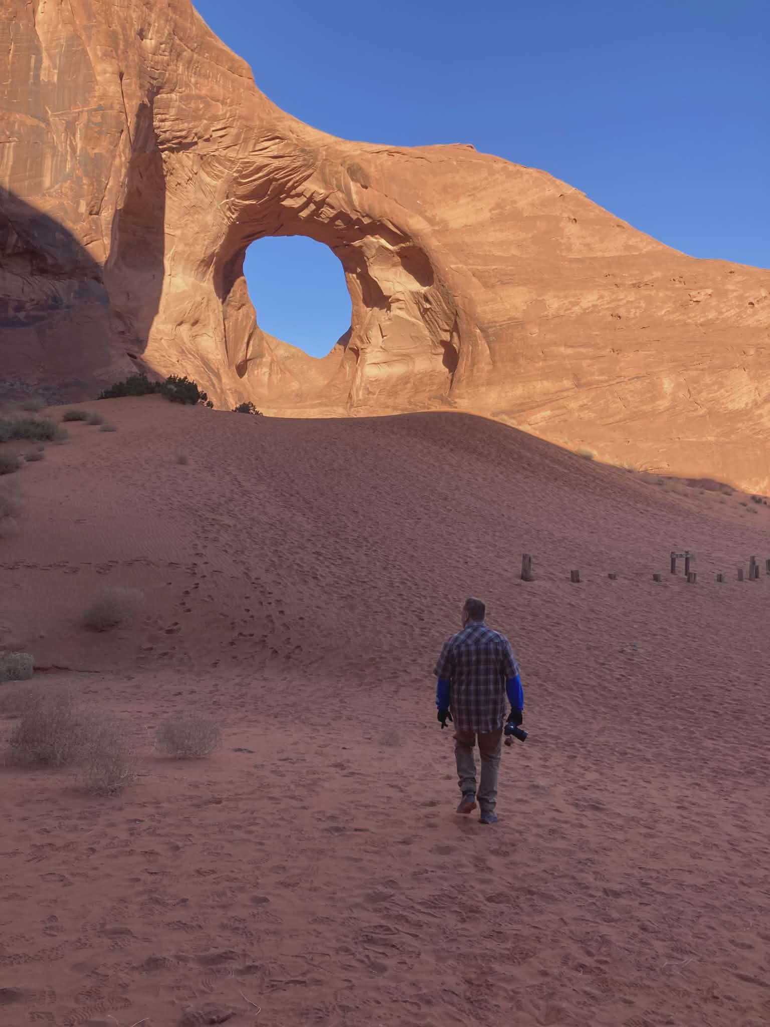 A man walking toward a large natural arch in a desert landscape with sandy ground and sparse vegetation, with a clear blue sky in the background.