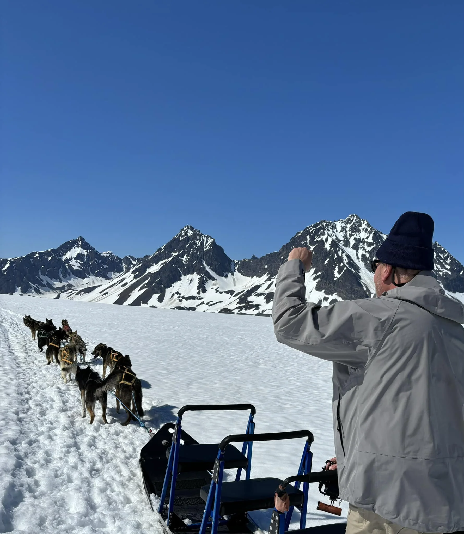 A man dressed in winter clothing and a beanie, standing next to a dog sled with multiple huskies, in a snowy landscape with tall mountain peaks under a clear blue sky.