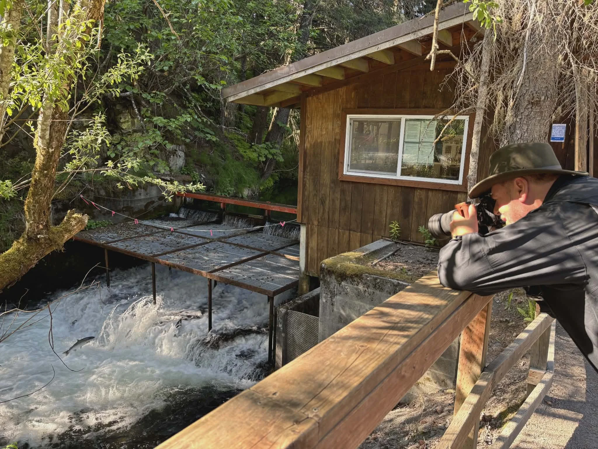 A man in outdoor gear taking photographs of a small waterfall and stream near a wooden cabin surrounded by trees and greenery.