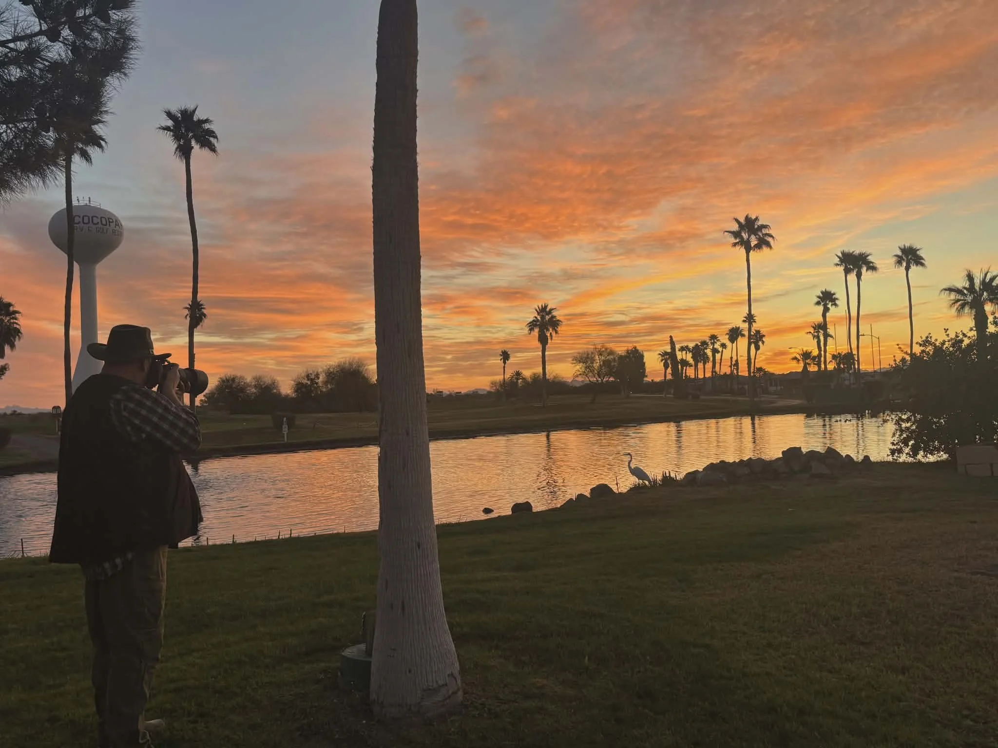 A person wearing a hat and dark clothing is taking a photo of a sunset over a lake, with palm trees silhouetted against an orange and pink sky, and a large water tower in the background.