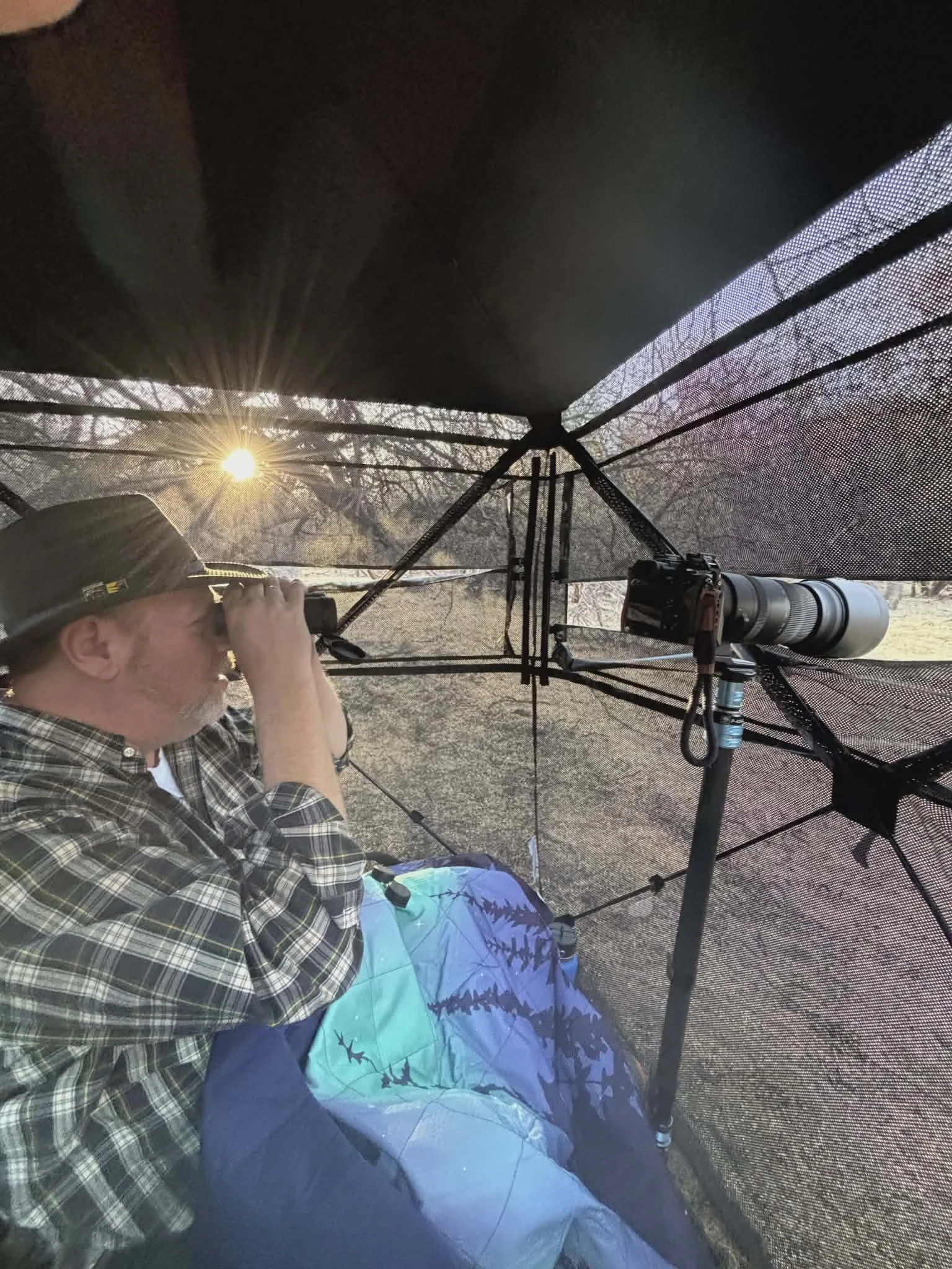 A man with a plaid shirt and hat sitting in a portable blind, looking through binoculars during sunset, with a telescope nearby.