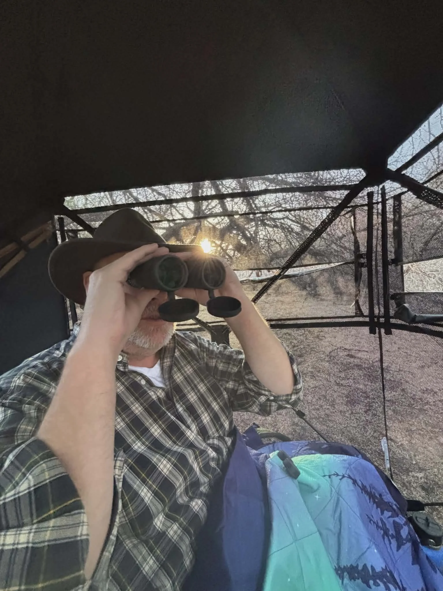 A man wearing a wide-brimmed hat and plaid shirt looking through binoculars inside a screened outdoor observation blind at sunset.