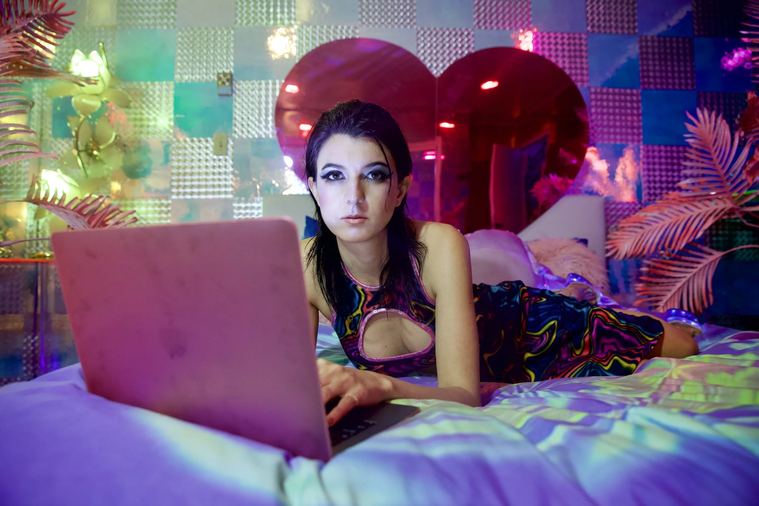 Author, Writer, Filmmaker, and Producer Isa Mazzei lays on a bed and types into a pink, plastic laptop. The colors are neon and bright.