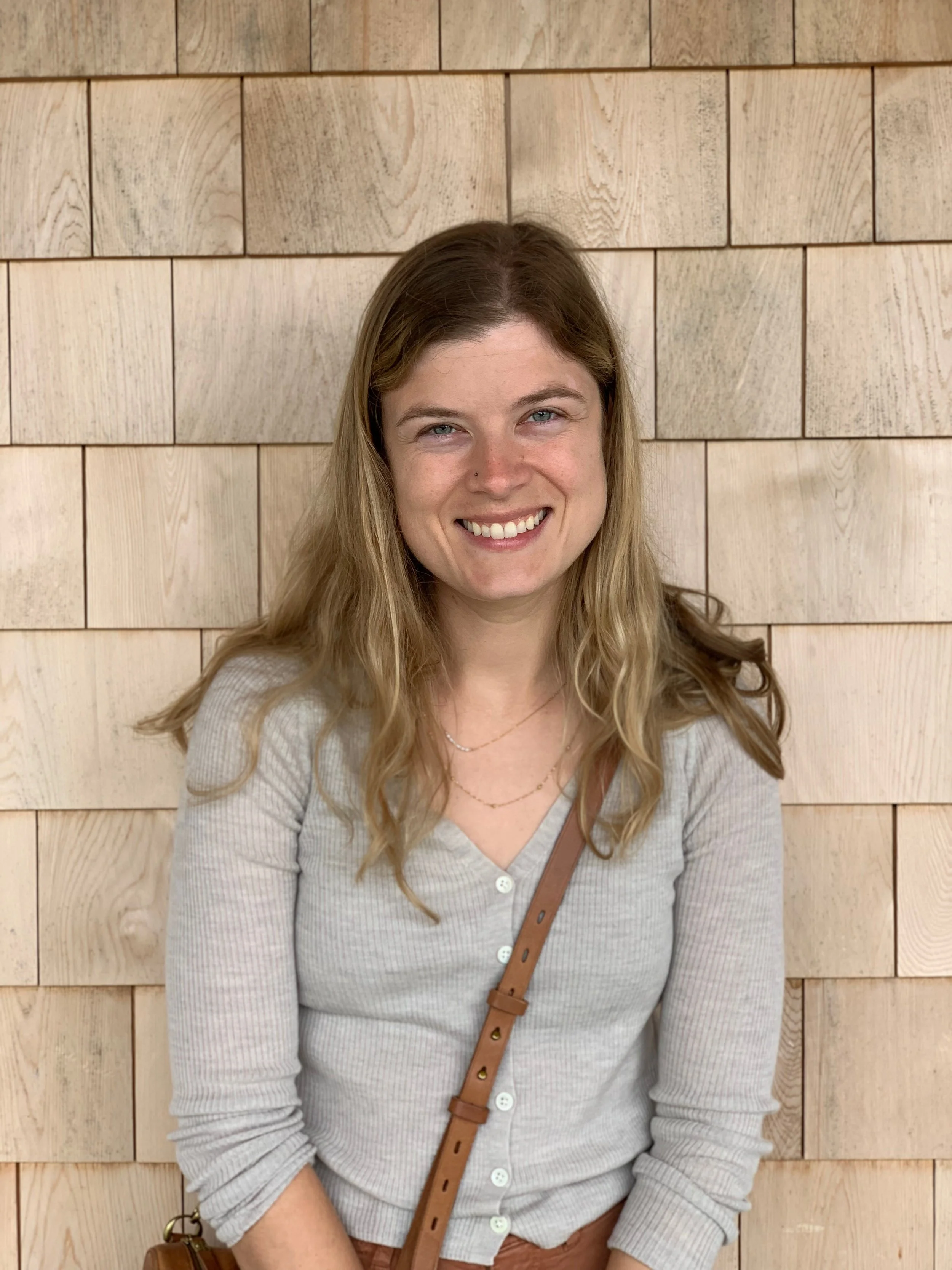 A young woman with blonde hair, smiling, standing in front of a wooden tile wall.