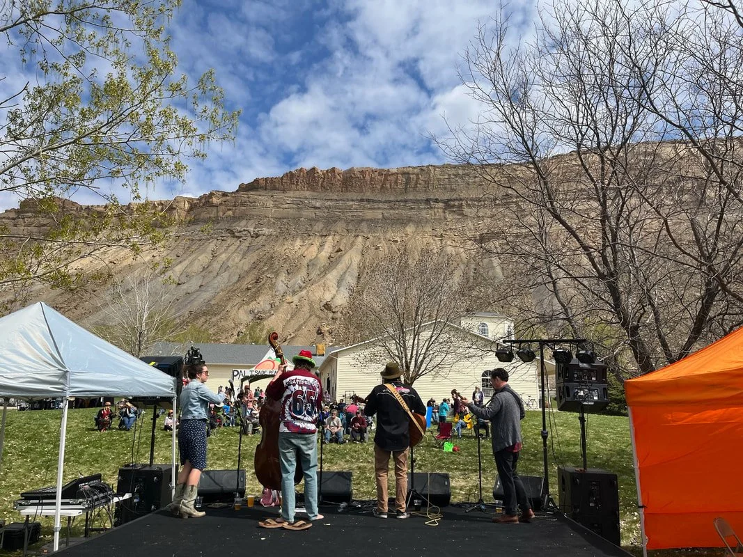 A live outdoor music performance on a small stage in front of a rock cliff with people sitting on the grass watching. There are tents on each side of the stage, and the sky is partly cloudy.