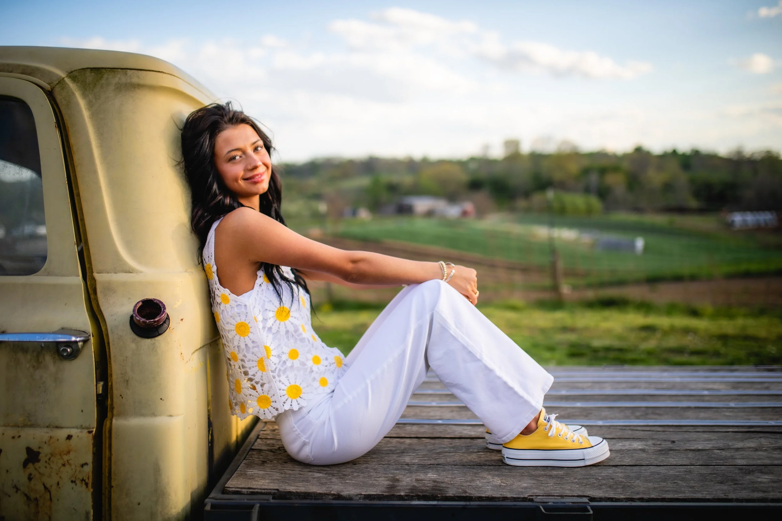 A woman sitting on the back of an old truck with a rural landscape in the background, wearing a white dress with yellow flowers and yellow sneakers, smiling at the camera.