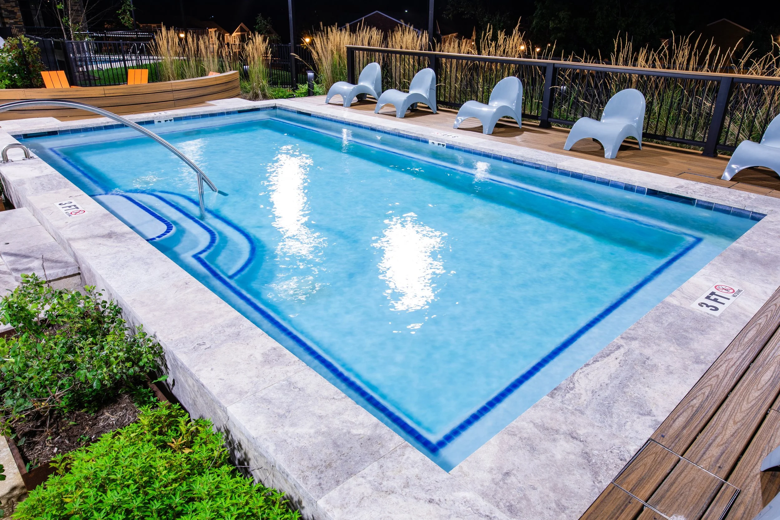 A nighttime view of a small rectangular outdoor swimming pool with a metal handrail, surrounded by a concrete deck. There are five modern white lounge chairs along the fence, which is adorned with tall ornamental grasses. The pool area is illuminated