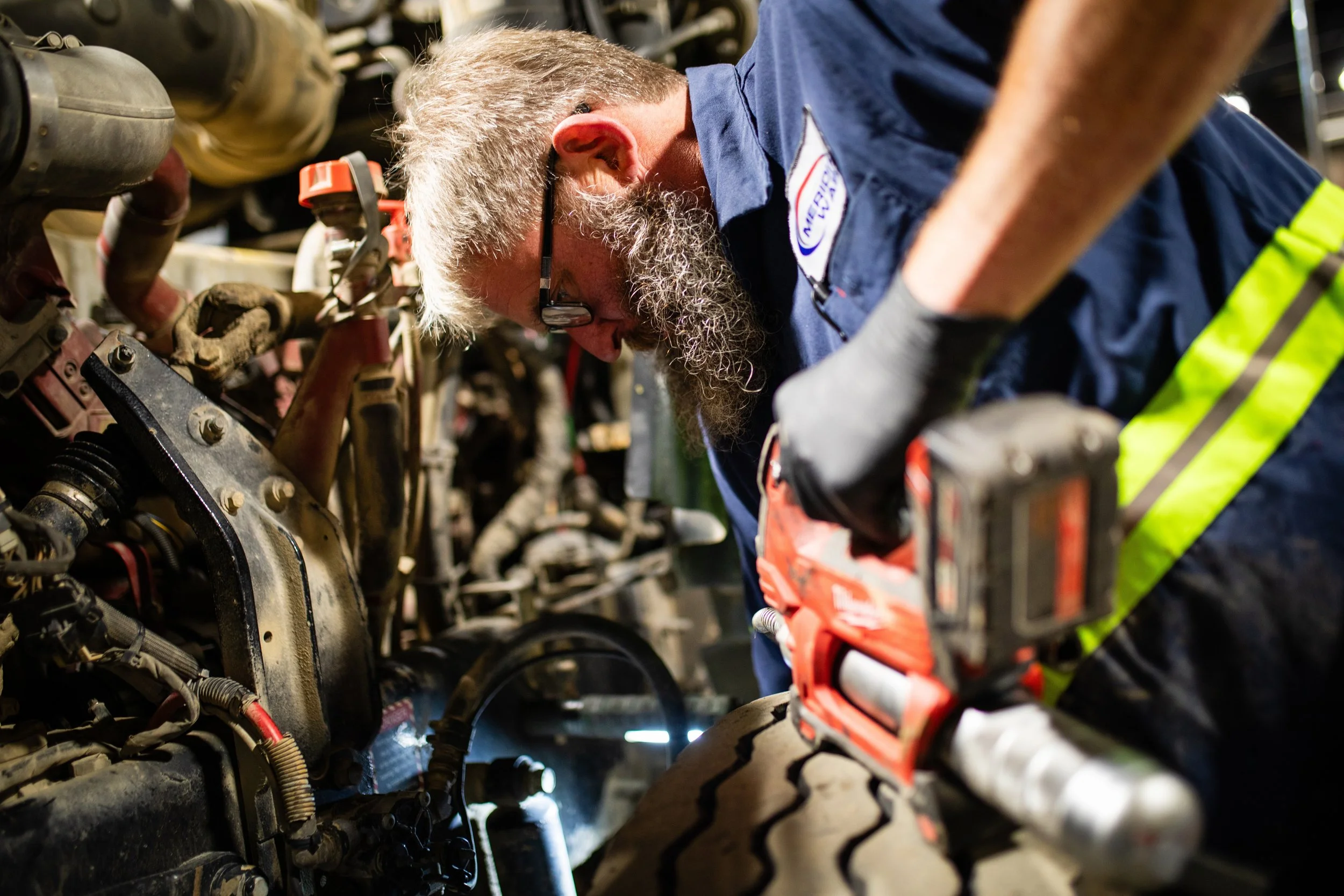 A mechanic with a beard, glasses, and a blue uniform is working on a vehicle engine with a power tool in a workshop.