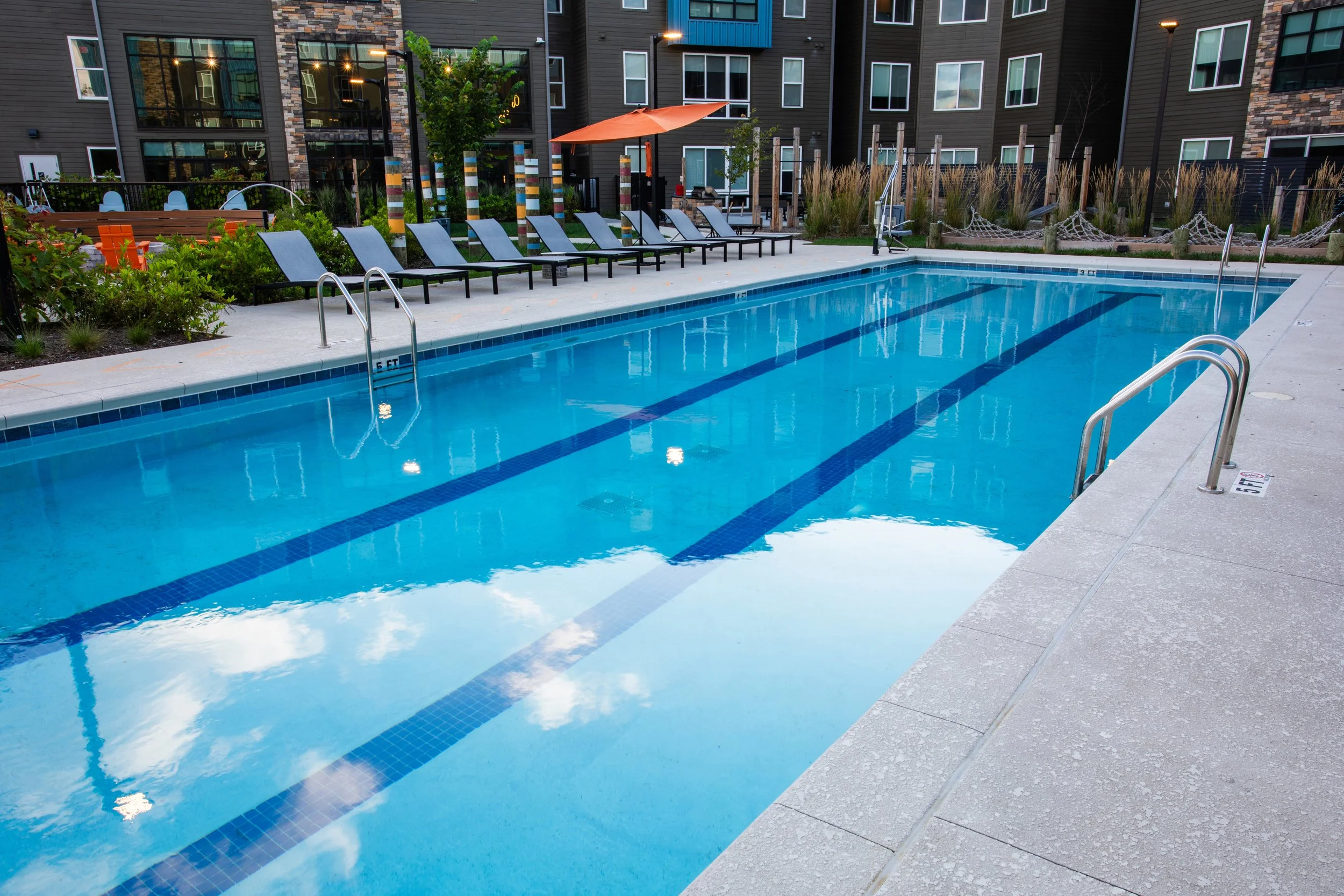 Empty outdoor swimming pool in a residential complex with poolside loungers and a shaded umbrella.
