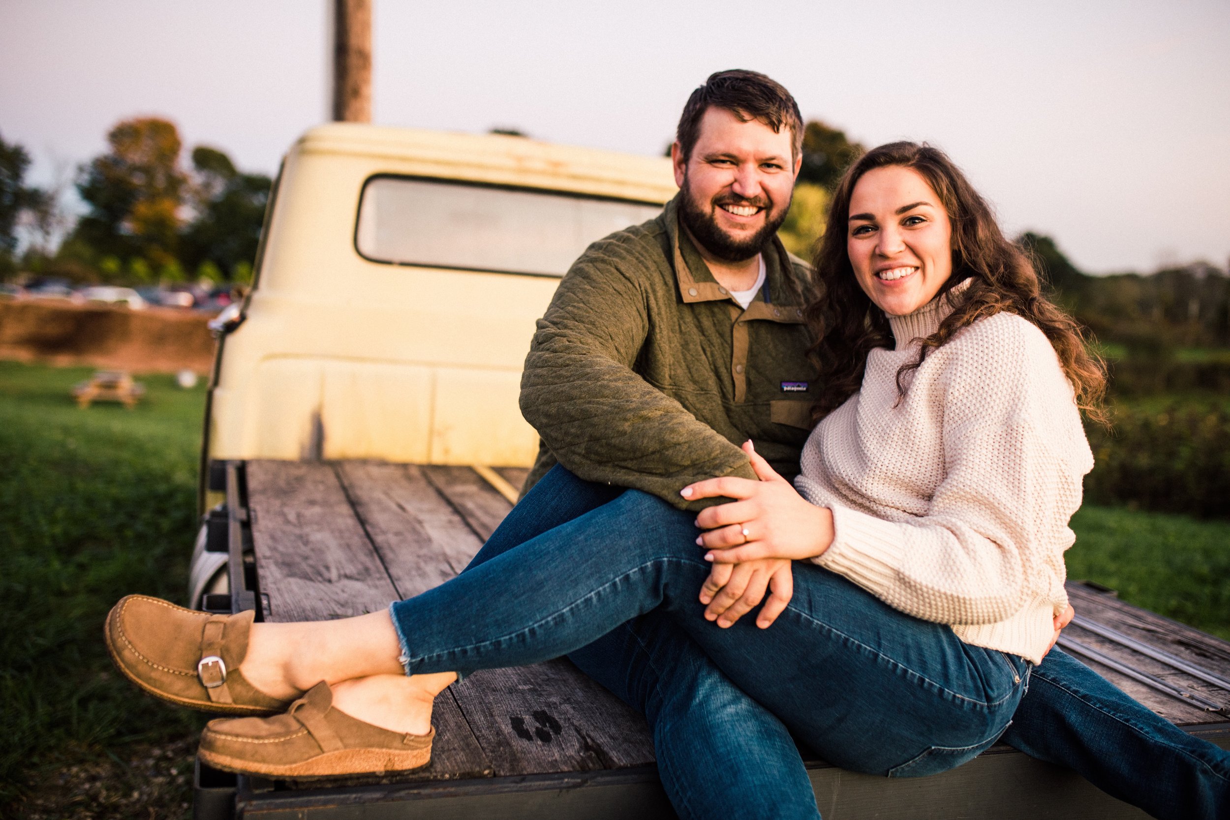A smiling couple is sitting on a wooden truck bed outdoors, with a grassy field and trees in the background. The man has a beard, and the woman has long, curly brown hair.
