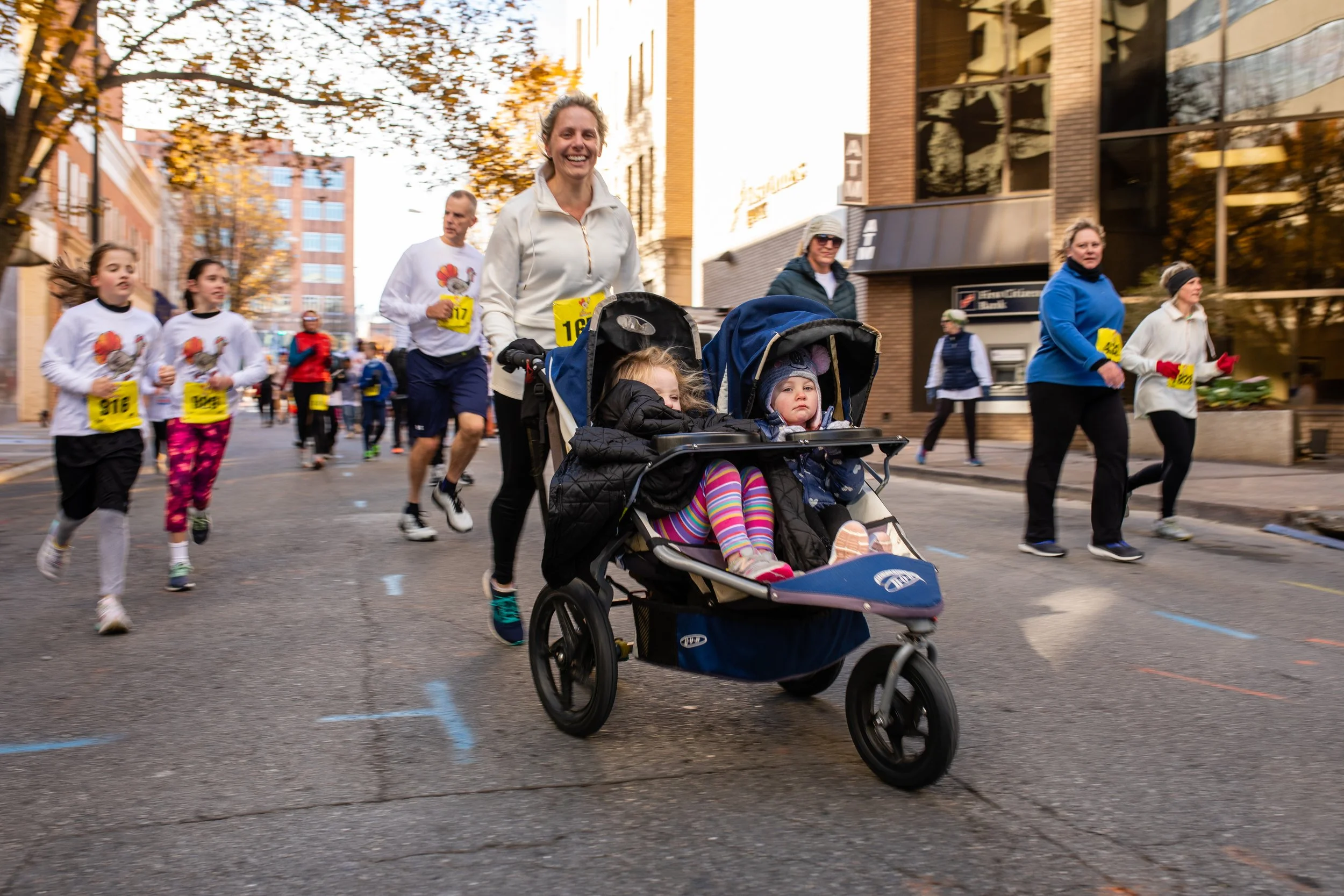 People participating in a charity race or marathon on a city street, including a woman pushing a stroller with two young children, some runners dressed in athletic wear, and community members, with autumn trees and buildings in the background.
