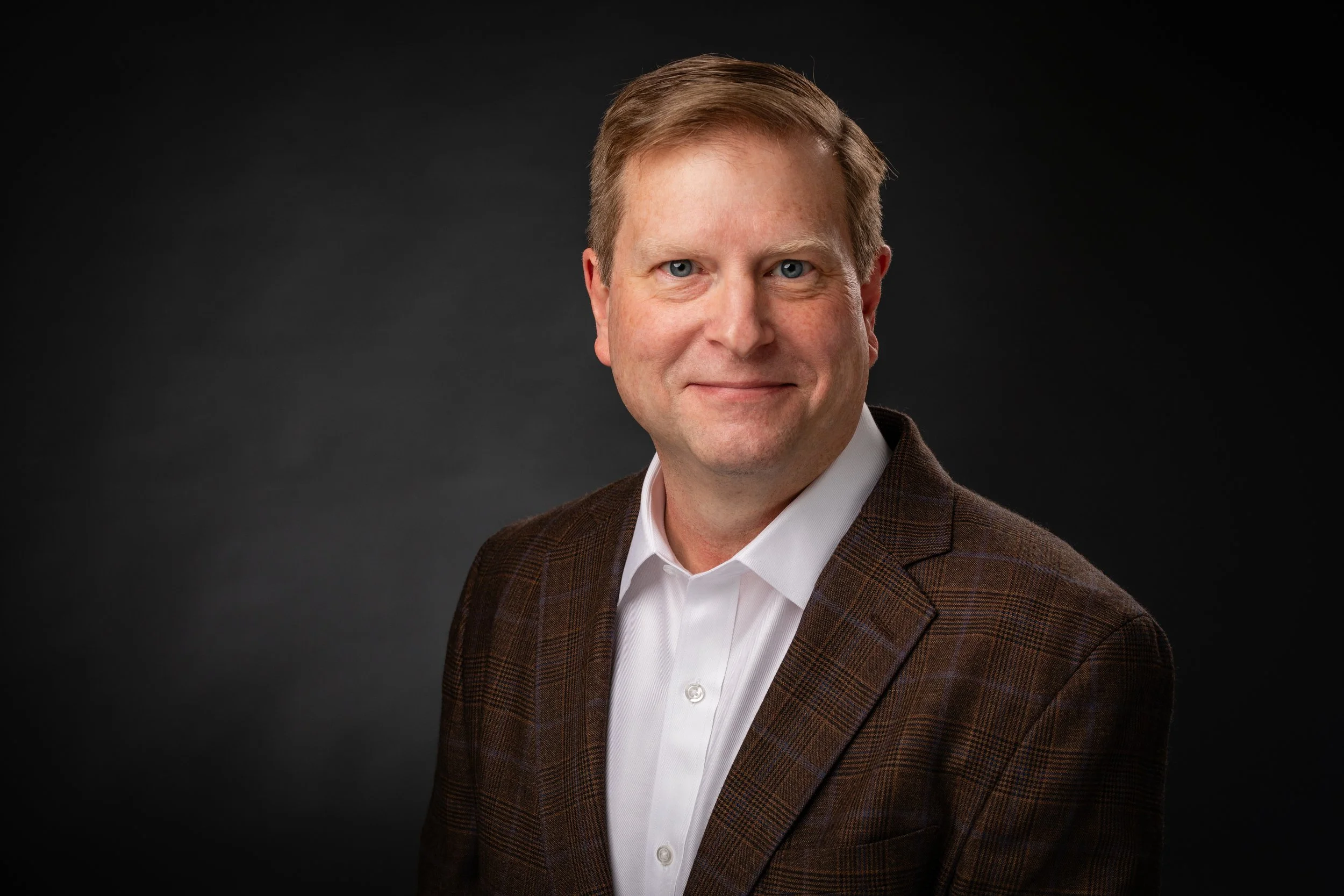Professional headshot of a man with short, light brown hair, wearing a white shirt and a brown plaid blazer, smiling against a dark background.