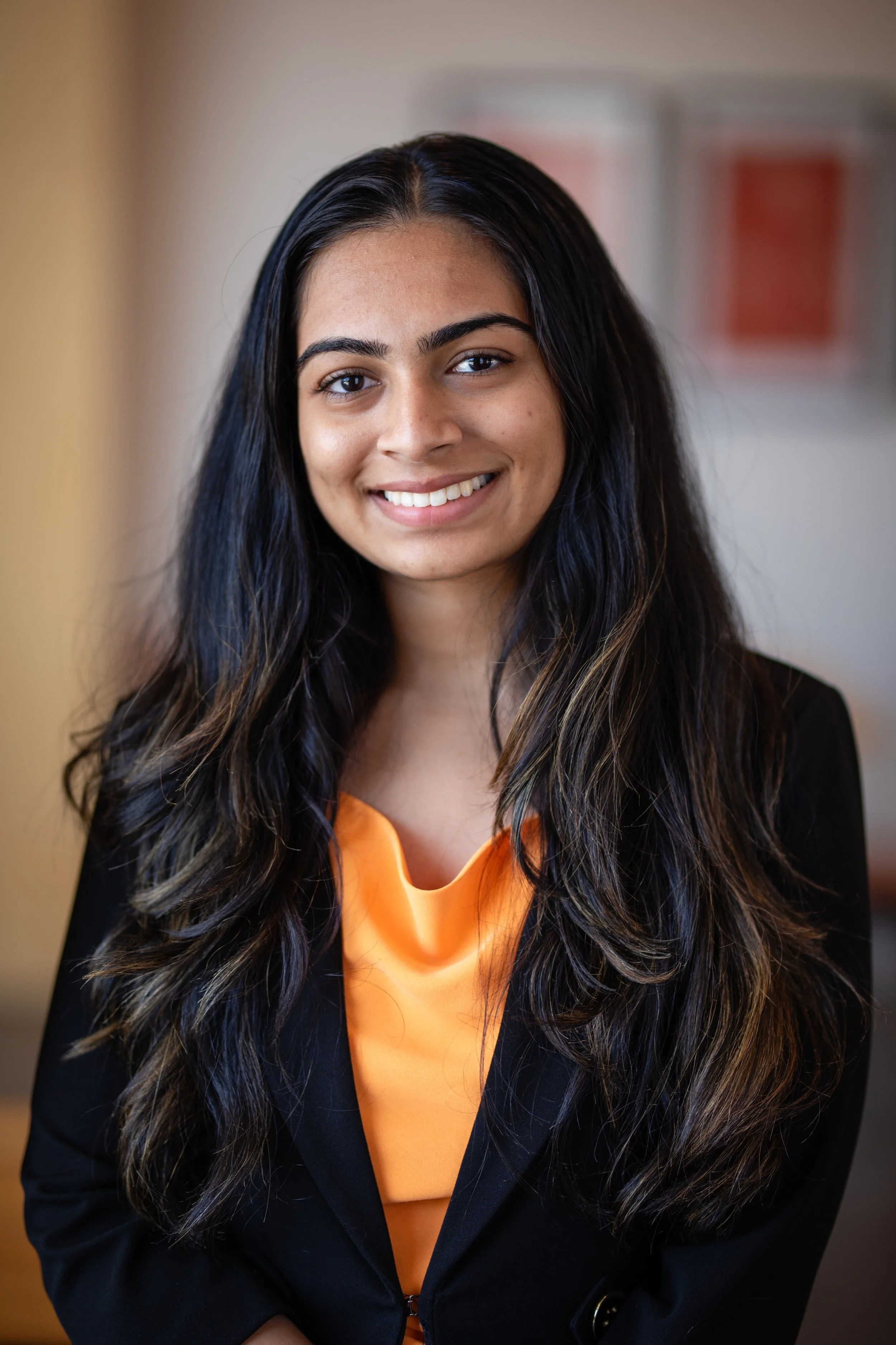 Portrait of a smiling woman with long dark wavy hair, wearing a black blazer over an orange top, inside a softly lit indoor setting.