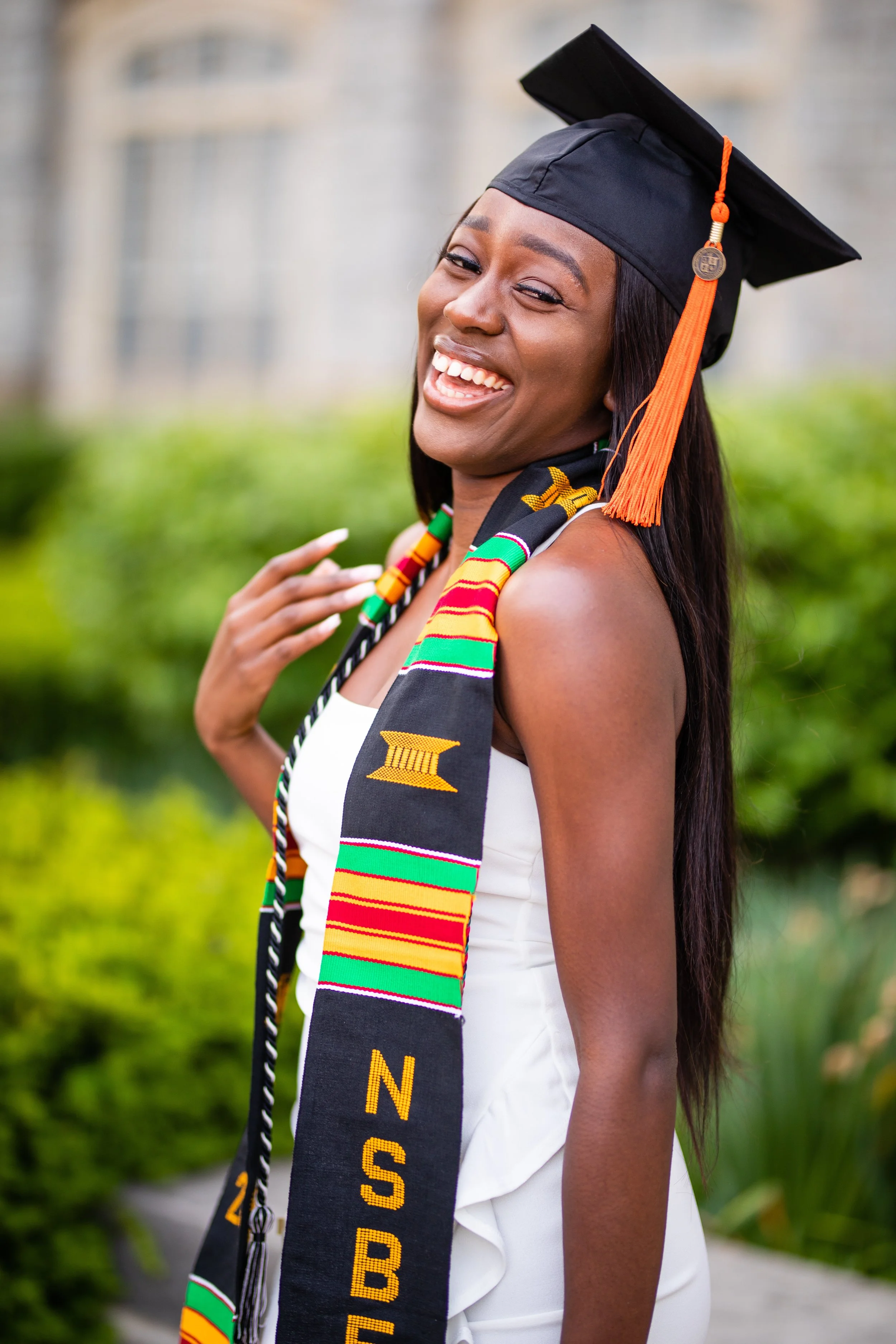 A young woman in a graduation cap and gown, smiling outdoors in front of greenery and a building, wearing a colorful stole.
