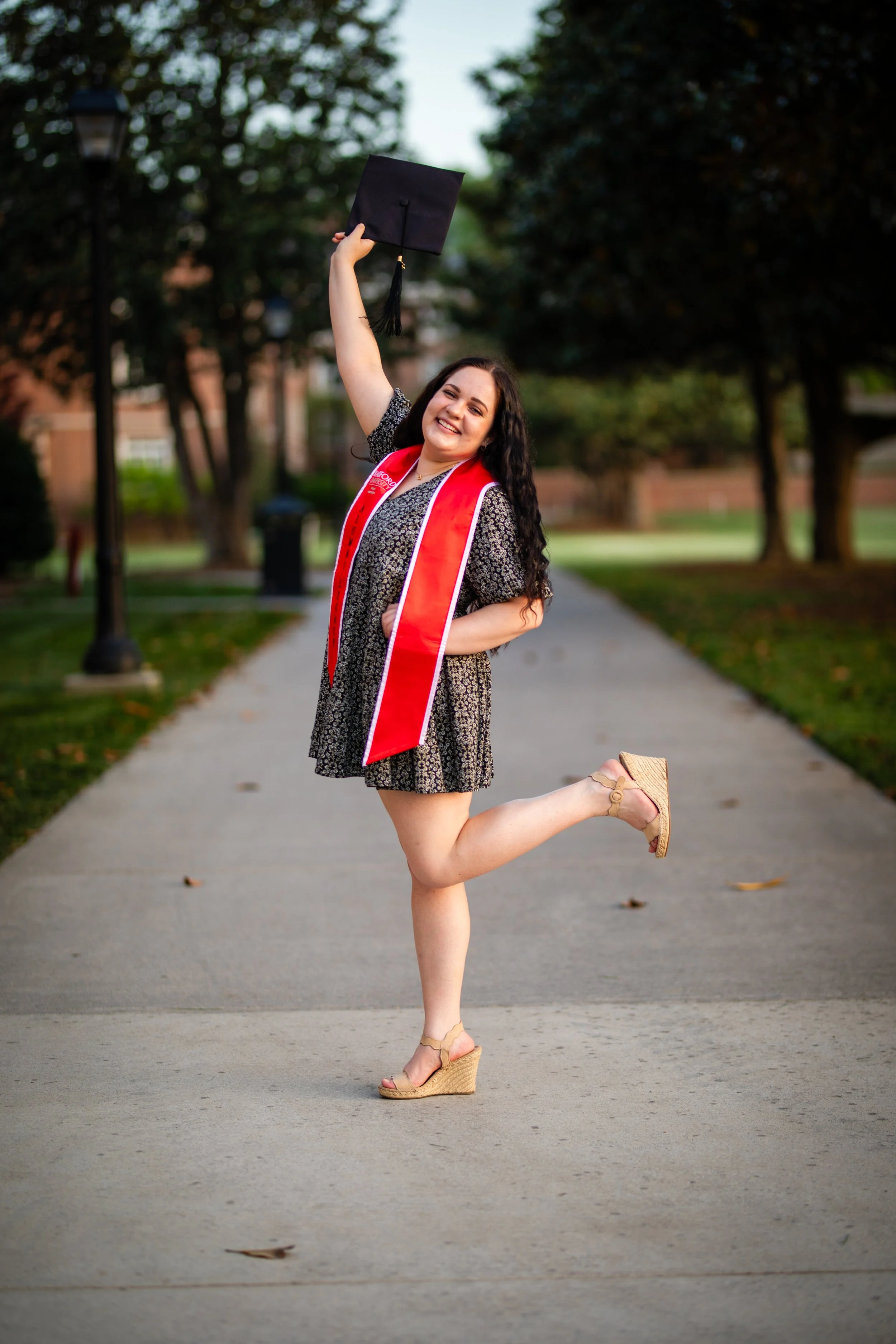 Woman celebrating graduation outdoors, holding a diploma in one hand and wearing a red stole, standing on a pathway surrounded by trees.
