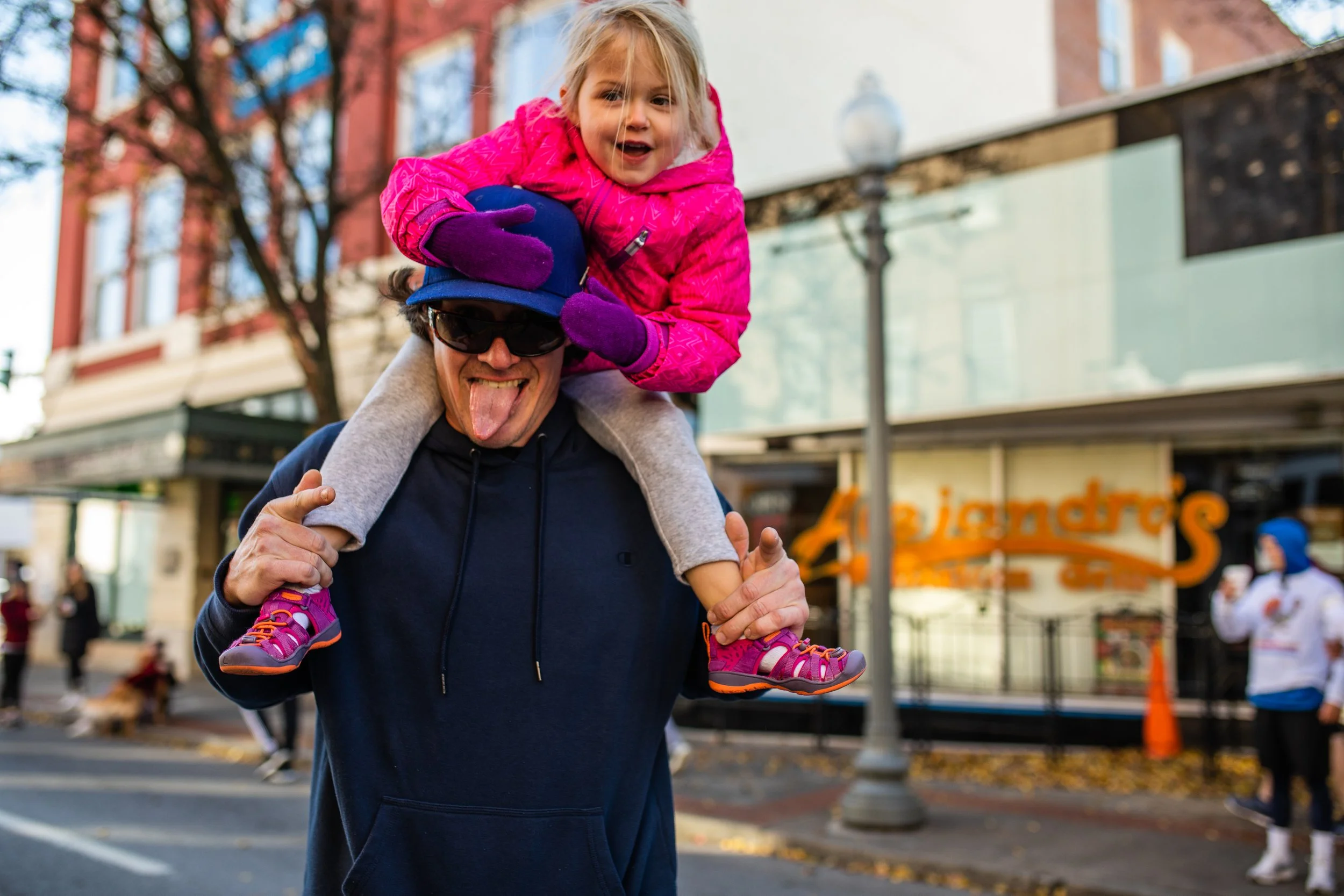 A man wearing sunglasses and a hoodie giving a piggyback ride to a young girl in a pink jacket and purple gloves on a city street, with a background of shops and pedestrians.
