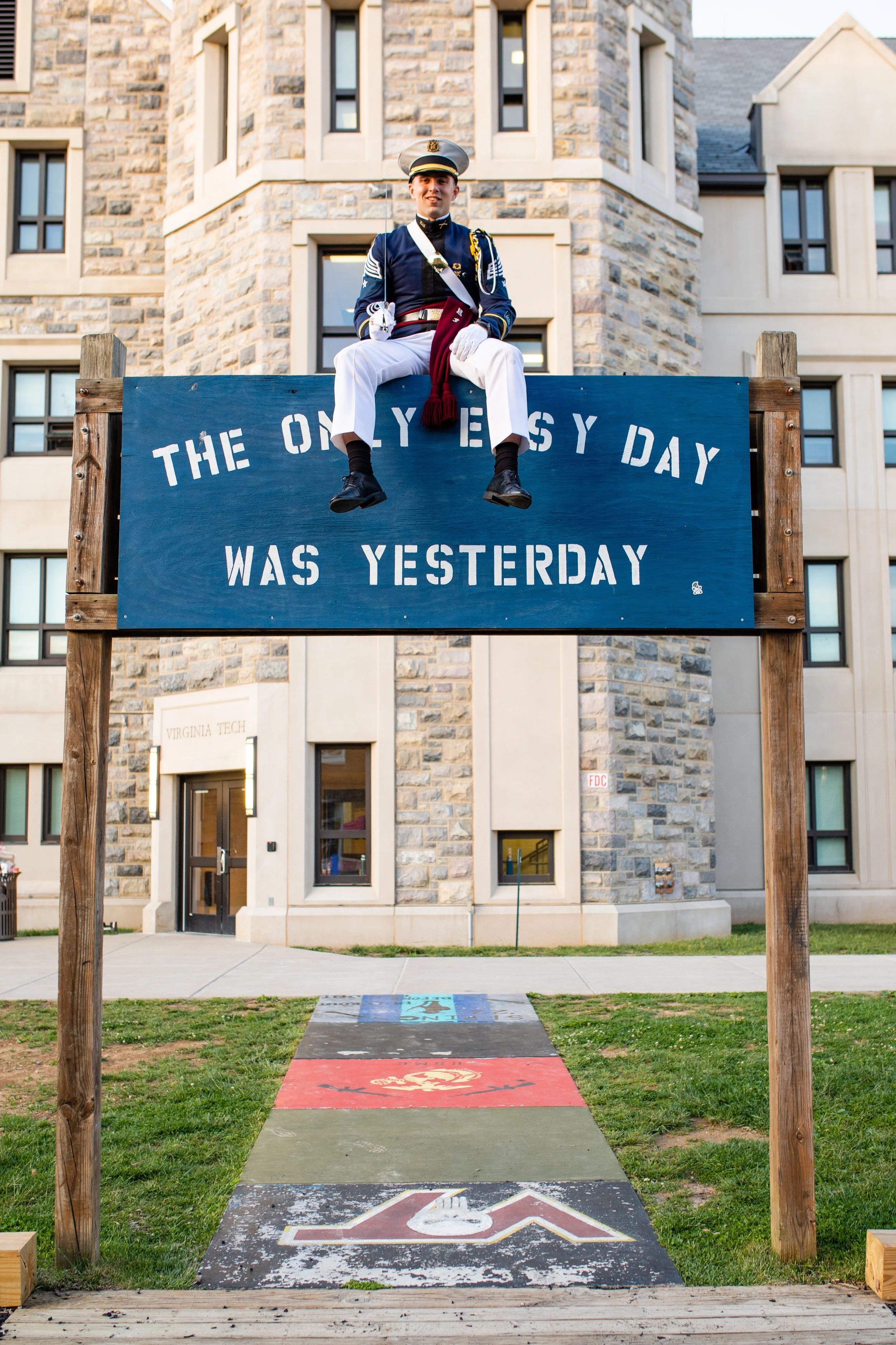 A person dressed in a military uniform sitting on a large blue sign that reads, 'The only easy day was yesterday.' The sign is set up in front of a stone building with a sign that says 'Virginia Tech' near the entrance.