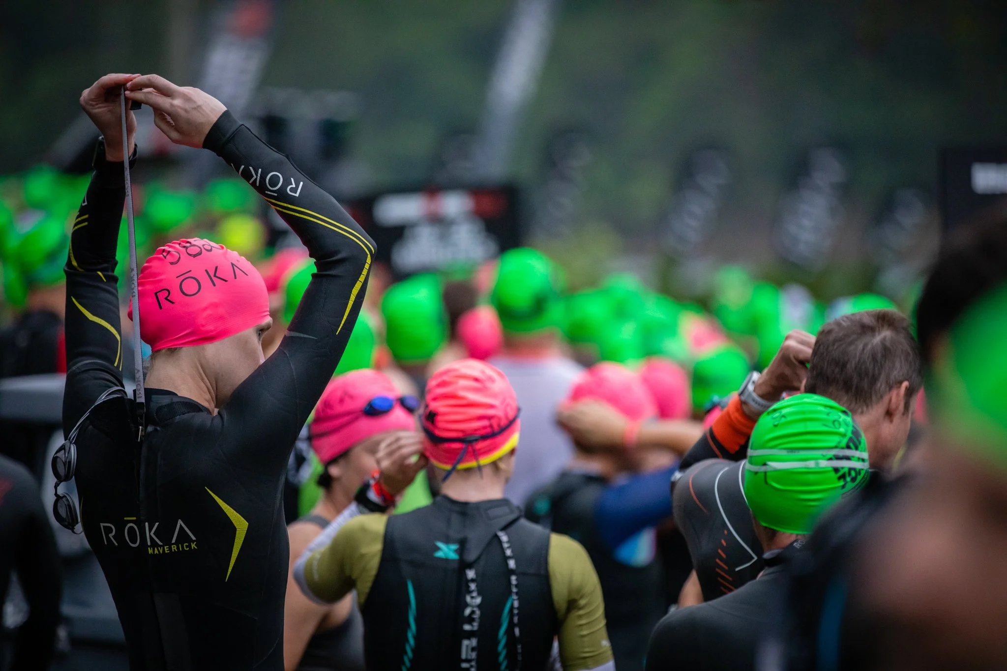 Triathletes wearing wetsuits and pink or green swim caps preparing for a race, with some adjusting goggles or clothing