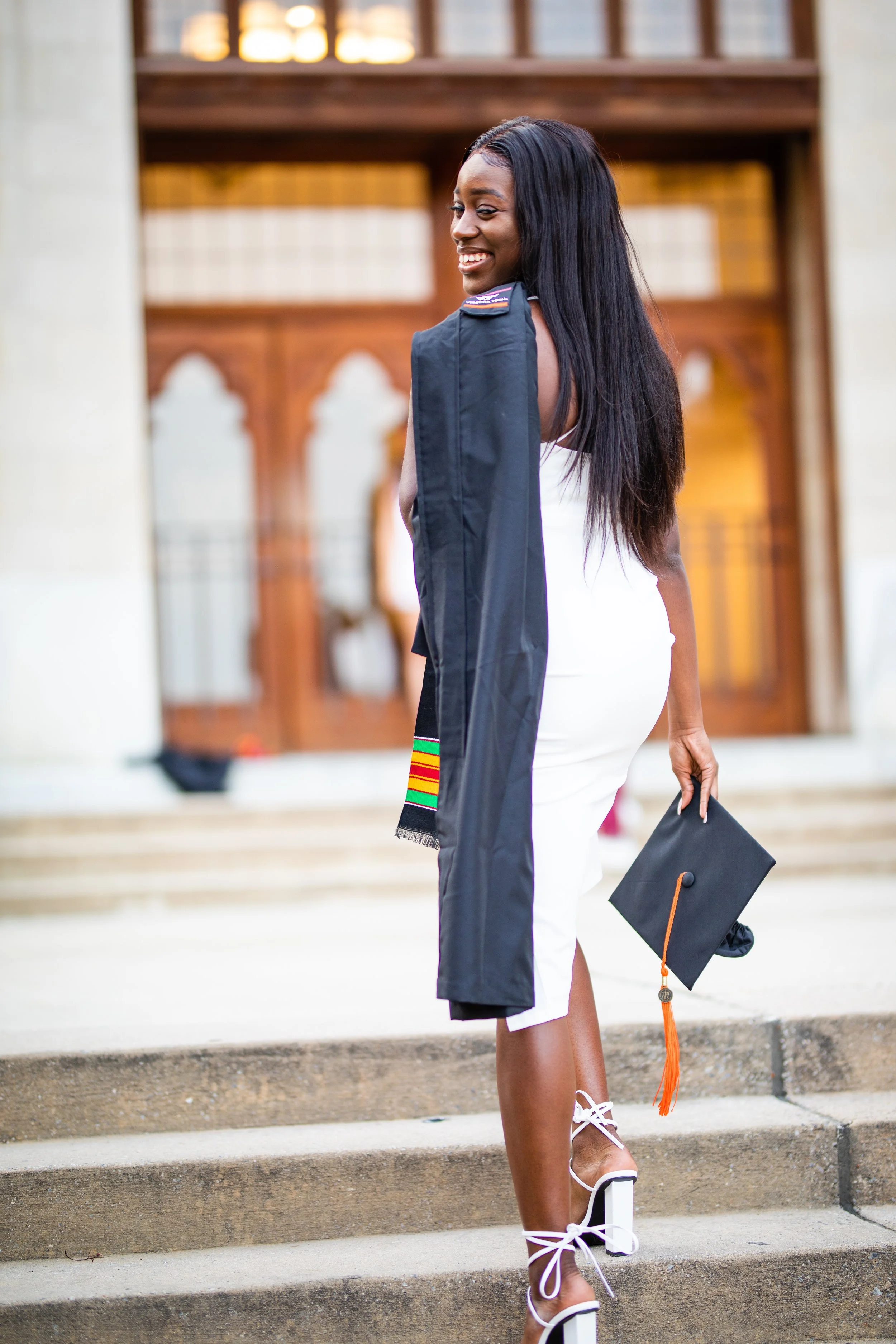 A woman in a white dress and high-heeled, lace-up sandals holding a graduation cap with an orange tassel, smiling as she stands on stone steps outside a building.