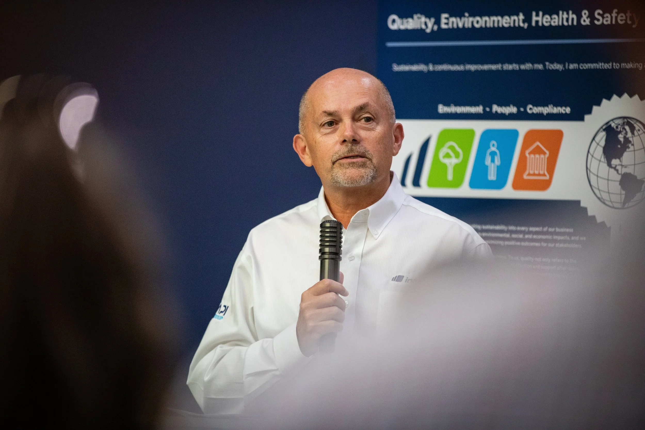 A man holding a microphone giving a presentation in front of a blue poster with environmentally related icons and the words 'Quality, Environment, Health & Safety'.