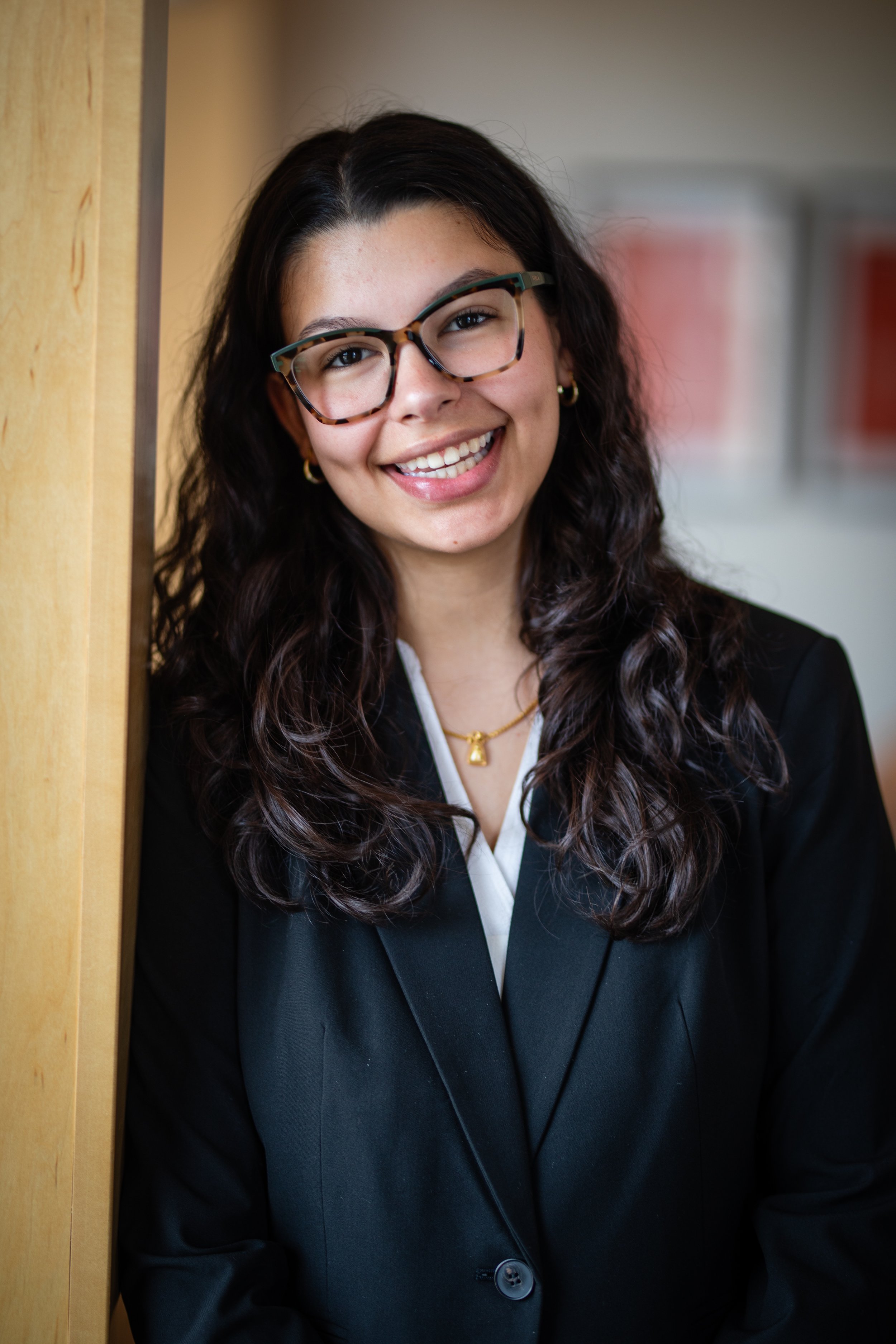 A young woman with long, curly dark hair, glasses, and light skin, smiling and leaning against a wooden partition in an indoor setting.