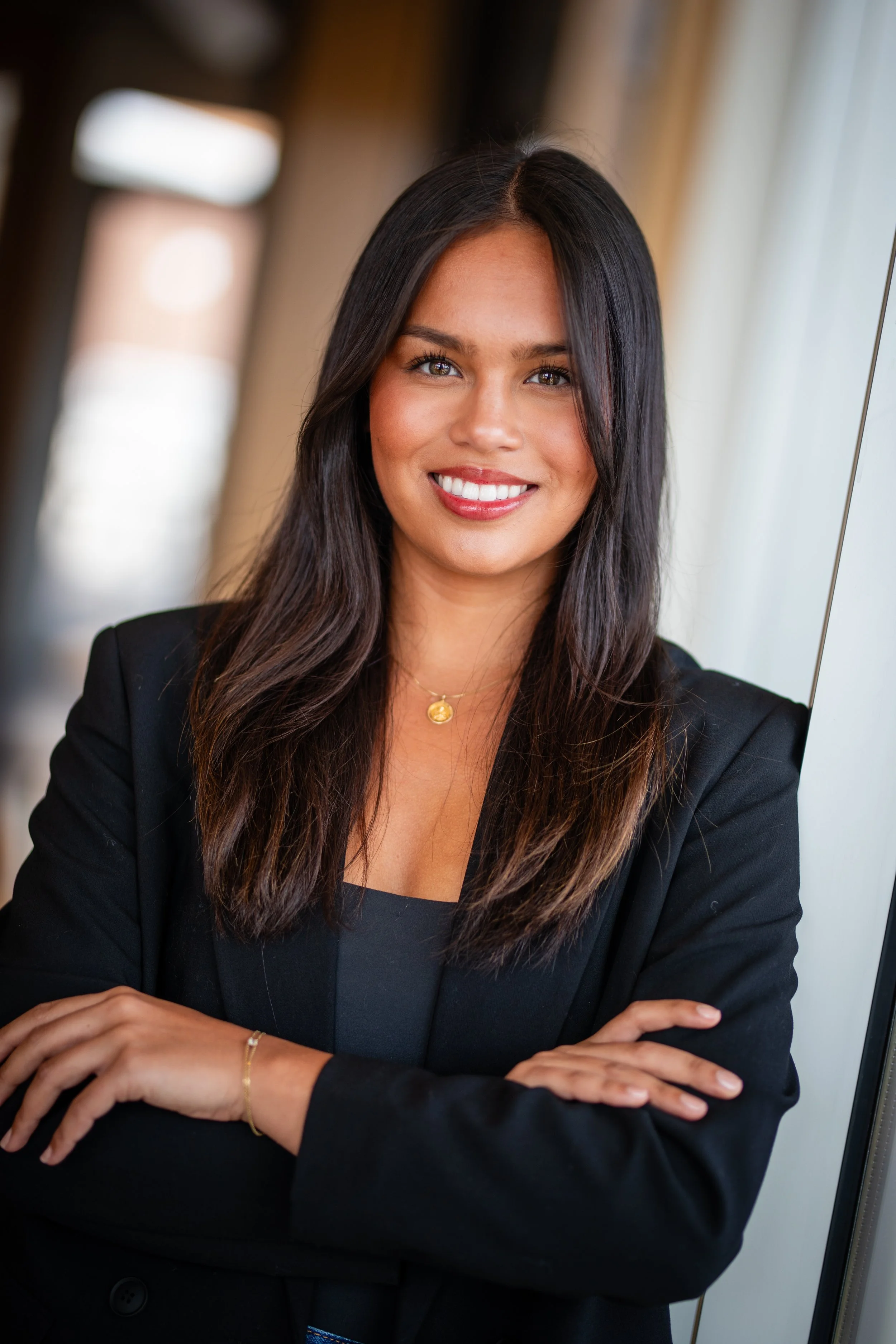 A woman with long dark hair, wearing a black blazer and black top, smiling and standing with her arms crossed near a window.