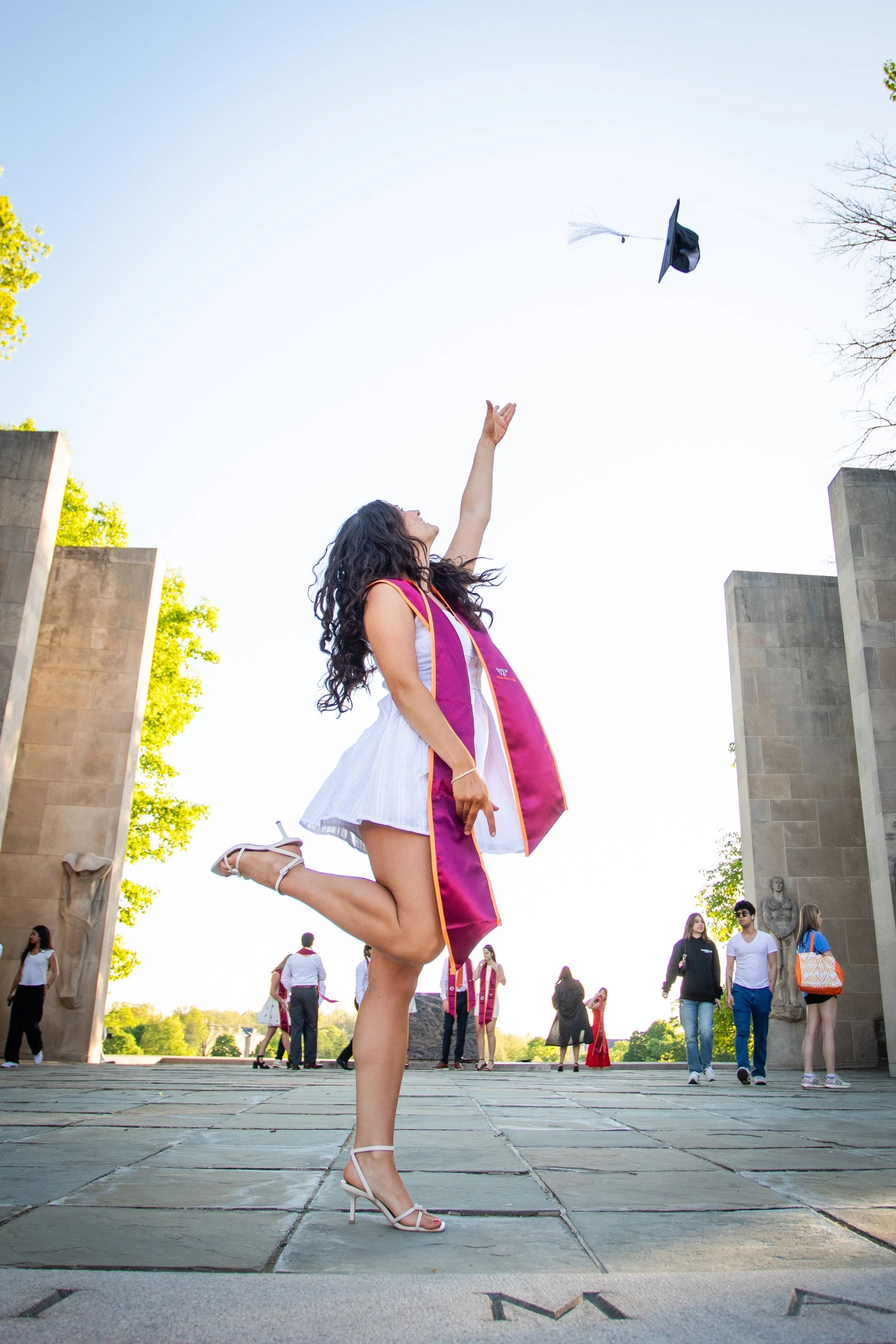 A woman in a graduation gown throws her mortarboard cap into the air during a graduation ceremony outdoors.