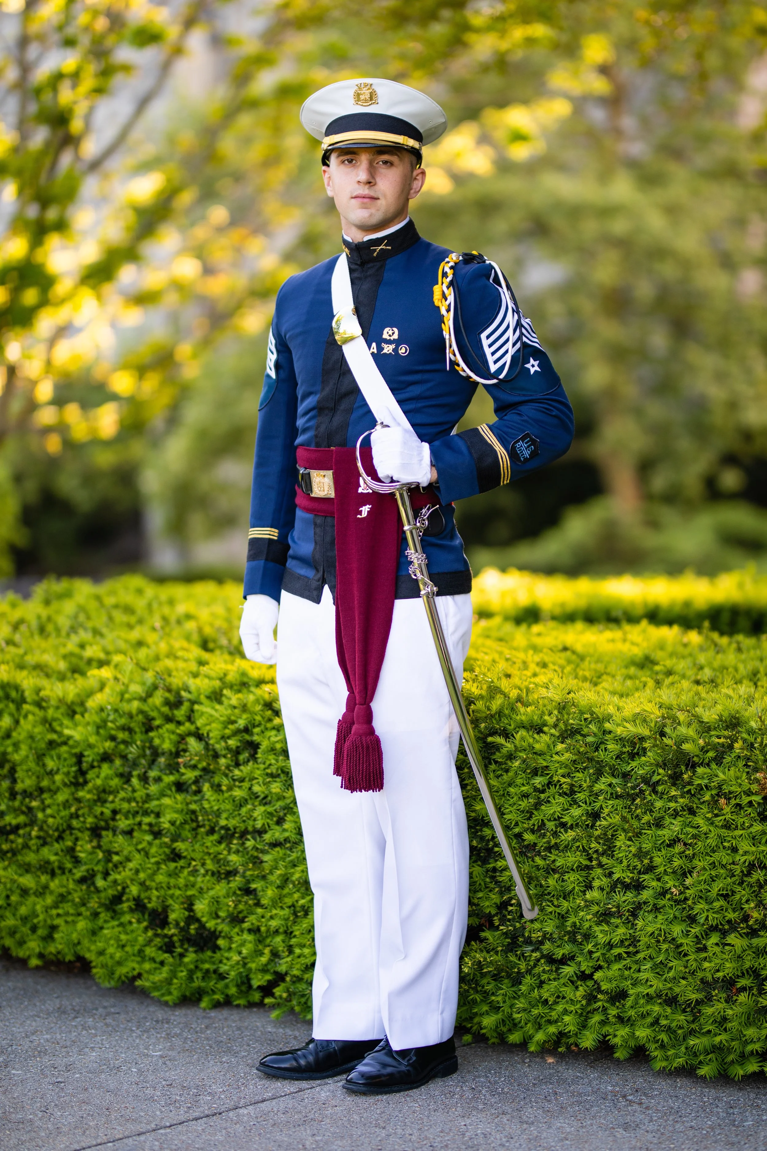 A young man dressed in a formal navy military uniform with white pants, black shoes, and a cap, standing outdoors in a garden with green bushes and trees in the background.