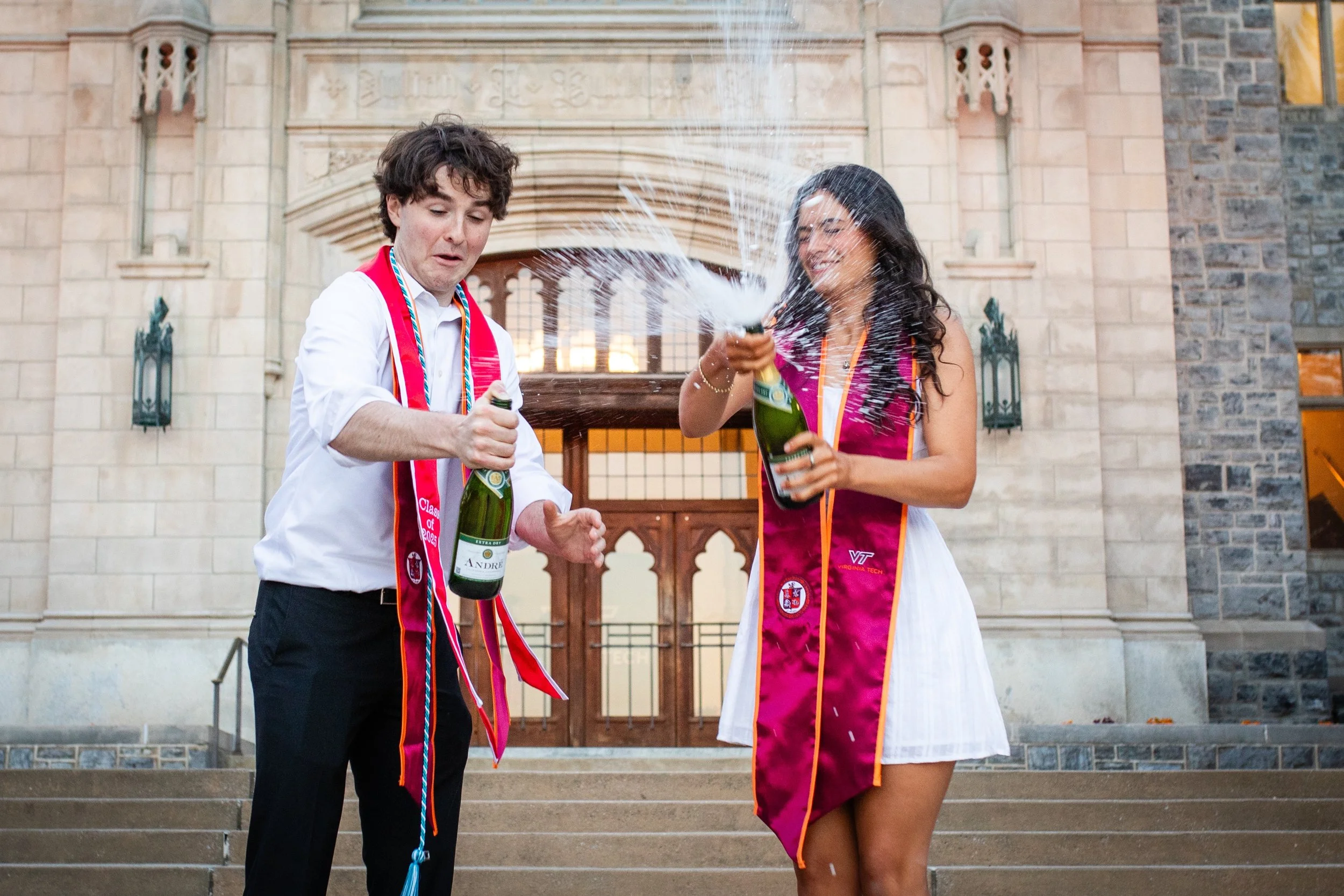 Two young adults, a man and a woman, celebrating together in academic regalia outside a building, popping champagne bottles and spraying champagne.