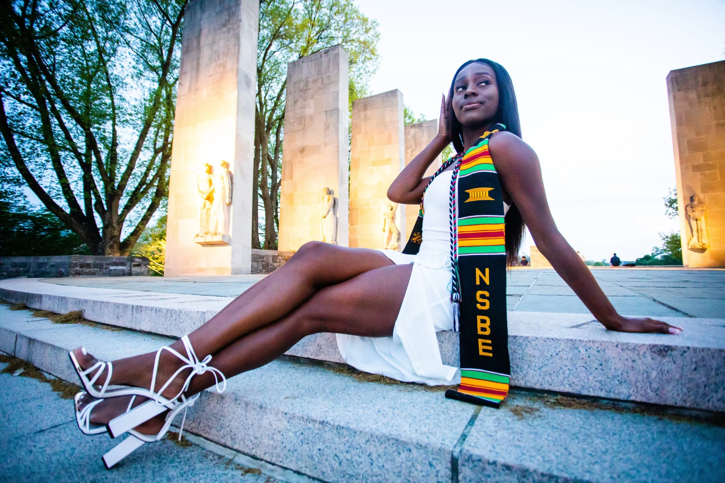 Young woman in a white dress and colorful graduation stole sitting on steps at a monument during dusk.