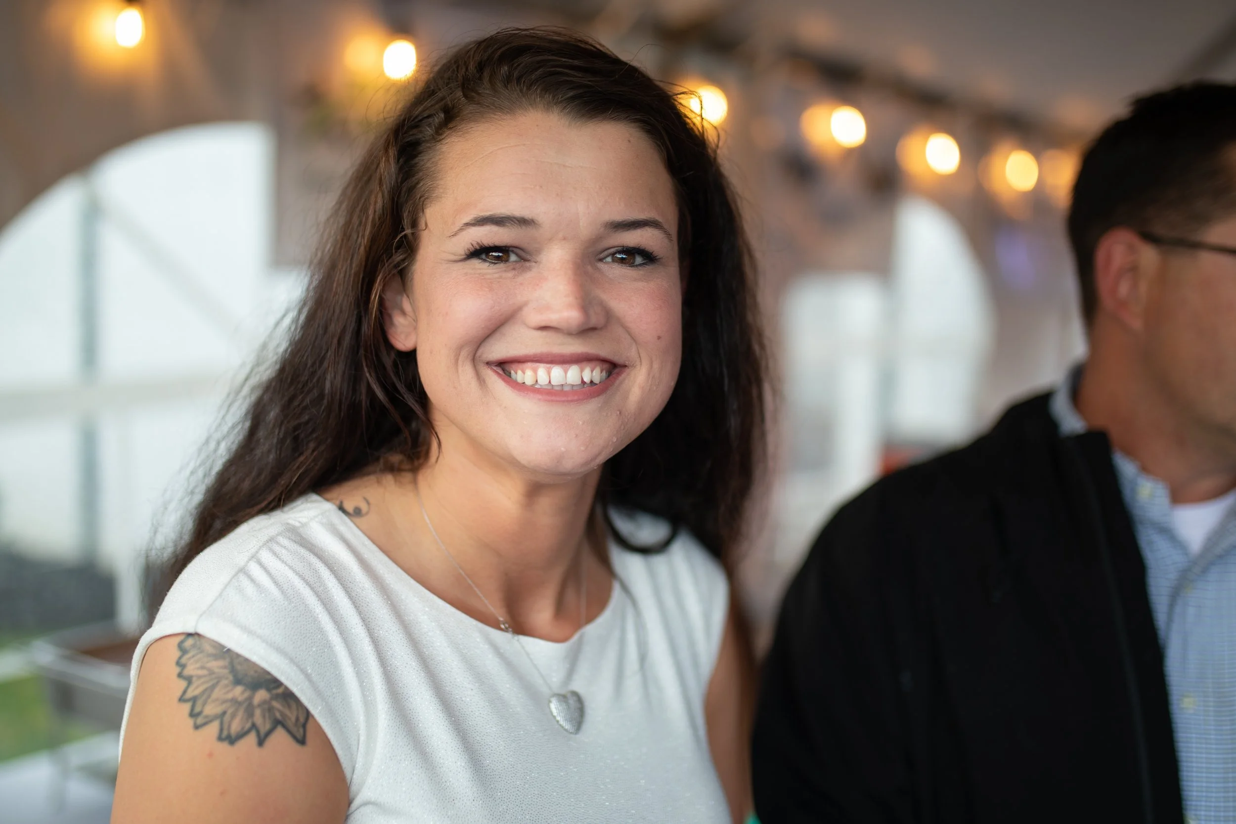 A woman with long dark hair smiling at the camera, wearing a white top with a tattoo on her right shoulder and a silver necklace with a heart pendant. In the background, there are blurred lights and windows, with part of a man visible on the right si