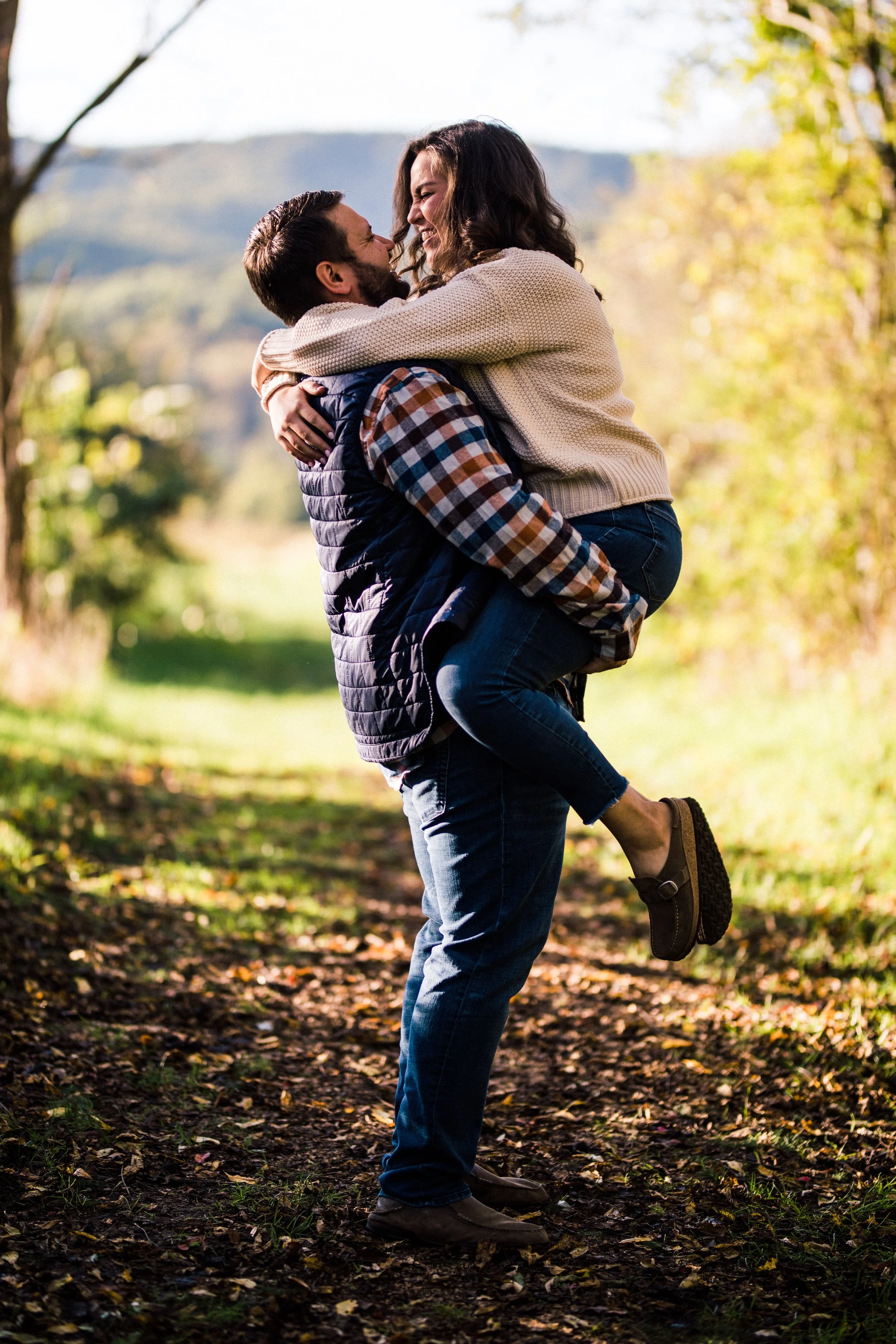 A man is carrying a woman on his shoulders in a sunlit outdoor setting with trees and a pathway, as they smile and laugh together.