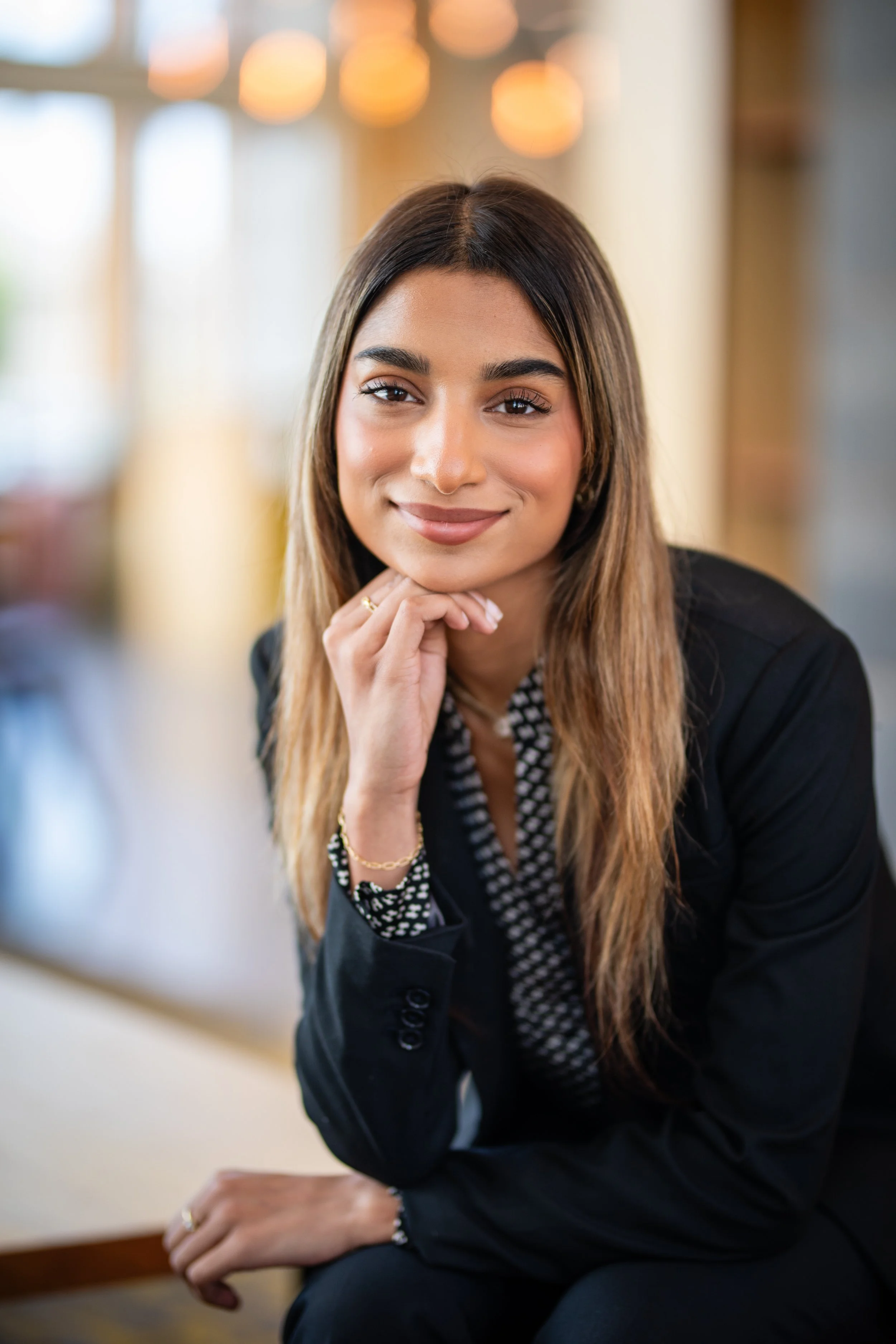 A young woman with long brown hair and a friendly smile, wearing a black blazer and patterned blouse, sitting indoors with a blurred background and warm lighting.