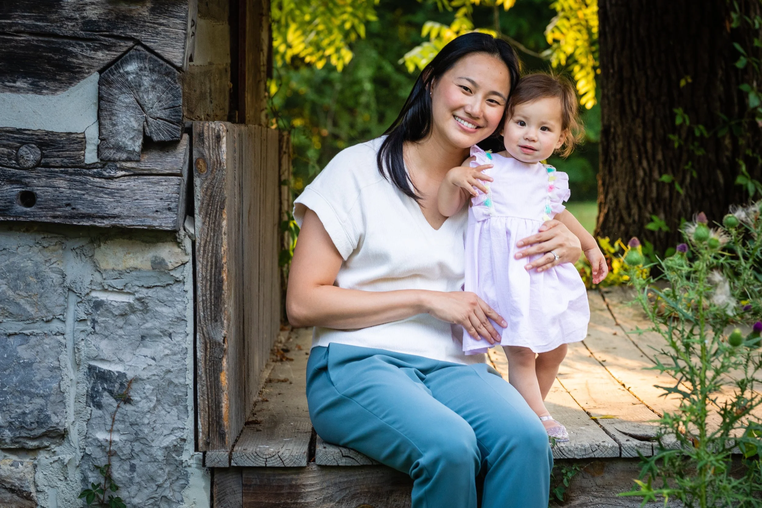 A woman and a young girl sitting on a rustic wooden bench outdoors, smiling and posing for the camera. The woman is wearing a white shirt and blue pants, while the girl is in a pink dress. There are trees and plants in the background.