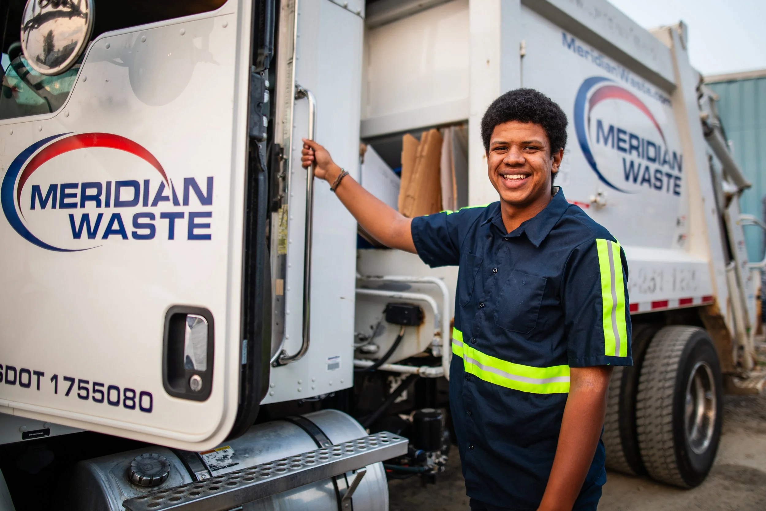 A smiling African American man in a navy blue uniform standing next to a white waste collection truck labeled 'Meridian Waste'.