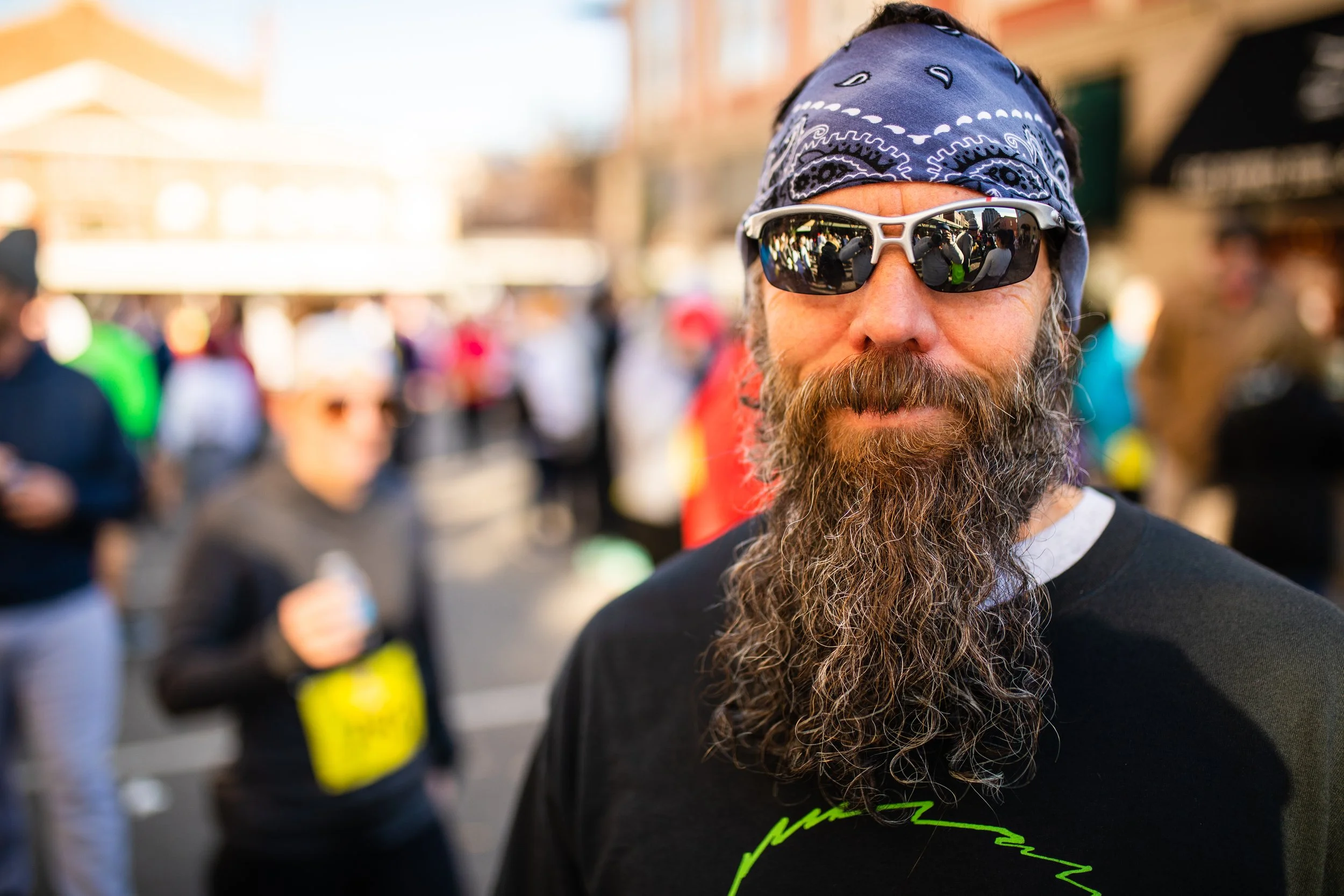A man with a long beard wearing sunglasses, a blue bandana, and a black shirt standing outdoors at a crowded event.