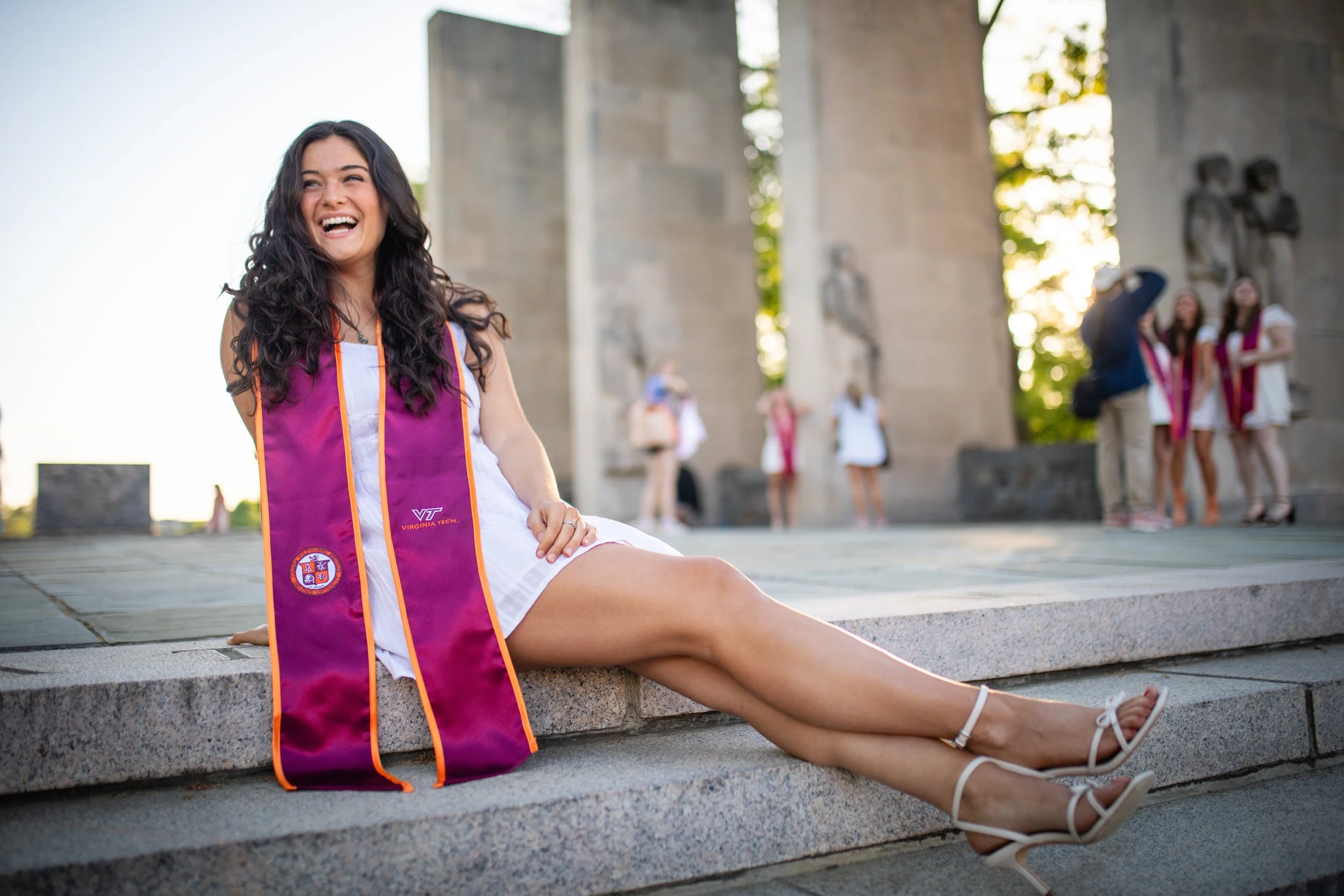 A woman in a white dress and white heels sitting on steps outdoors, wearing a pink and purple graduation stole with the Virginia Tech emblem, smiling and laughing, with other graduates in the background.