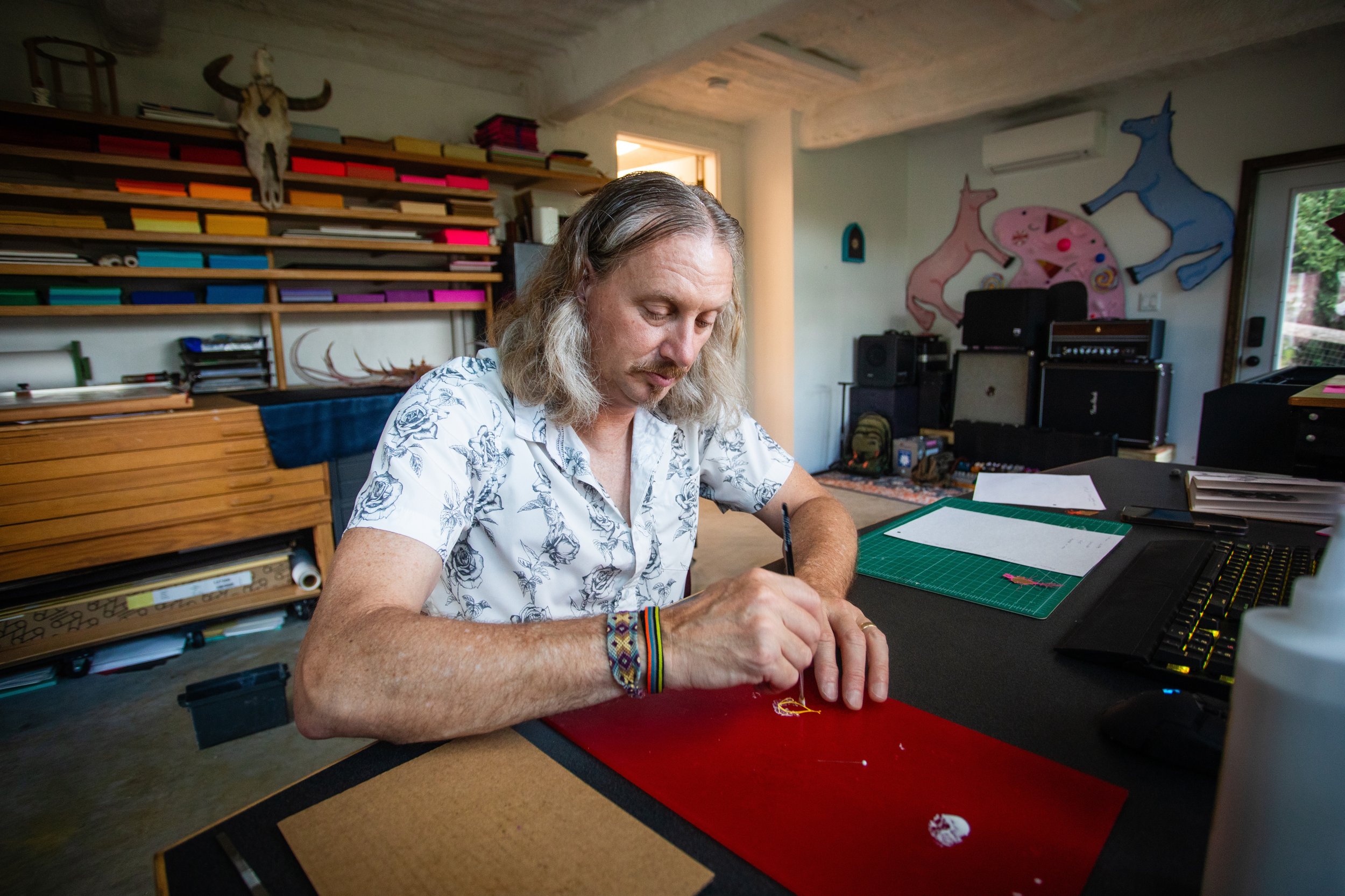 A man with long gray hair and a floral shirt is sitting at a desk, working on a craft project with a needle and thread in a room decorated with colorful animal artwork and musical equipment.