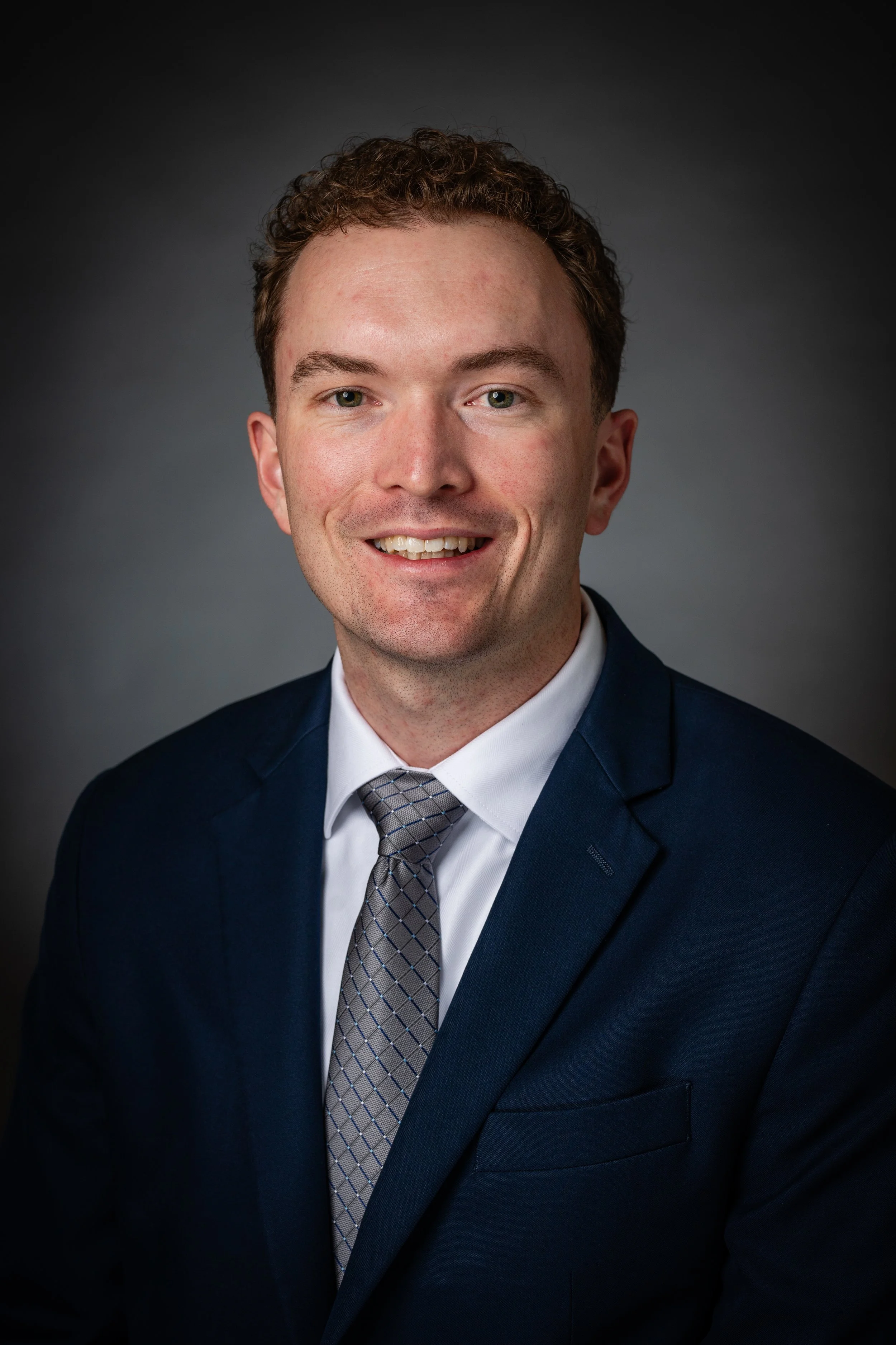 Professional headshot of a young man with curly hair, wearing a dark blue suit, white shirt, and patterned gray tie, smiling against a gray gradient background.