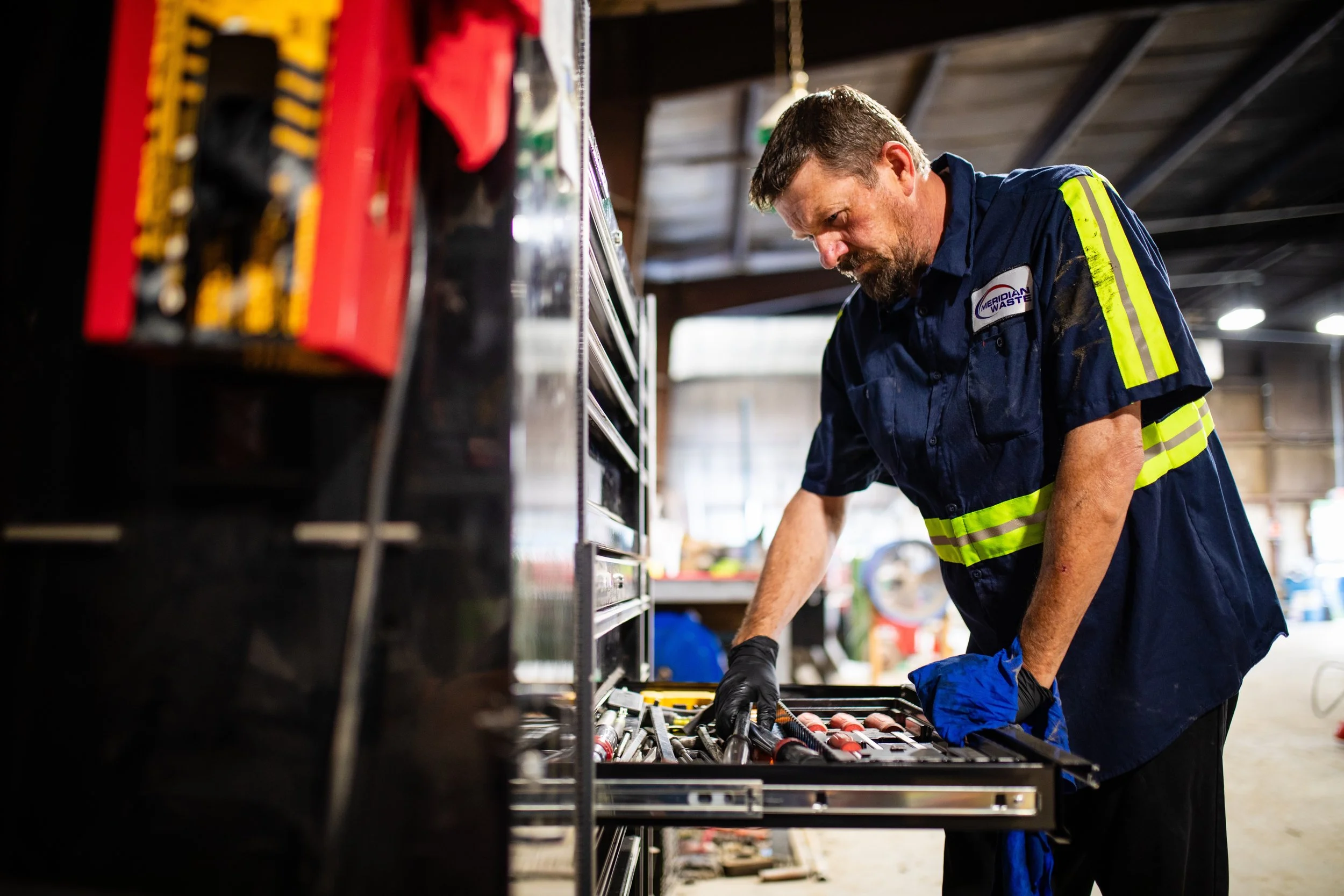 A male worker organizing tools in a workshop or garage, wearing a navy blue uniform with reflective yellow stripes and black gloves.