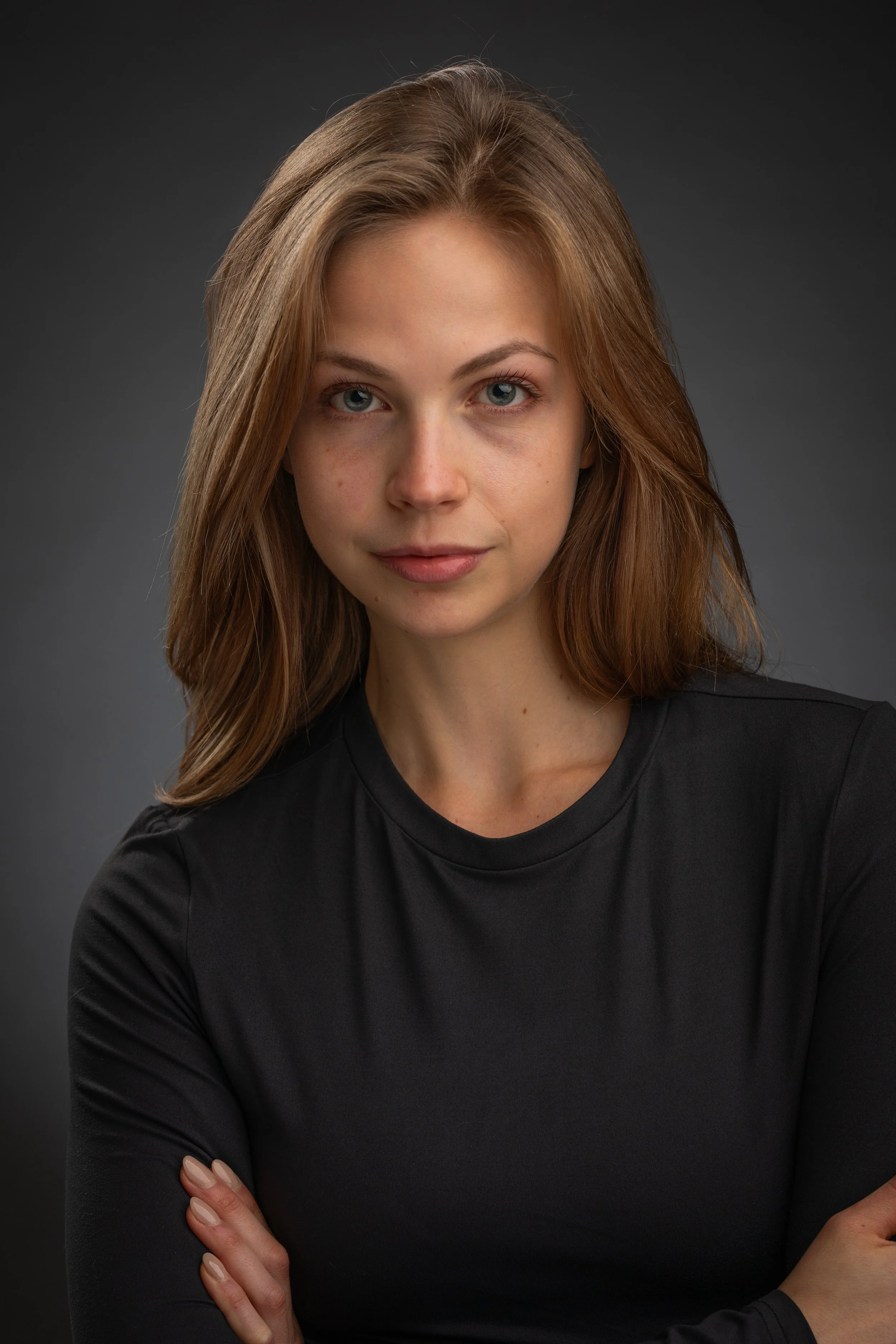 Portrait of a young woman with light brown hair and blue eyes wearing a black shirt against a dark gray background.