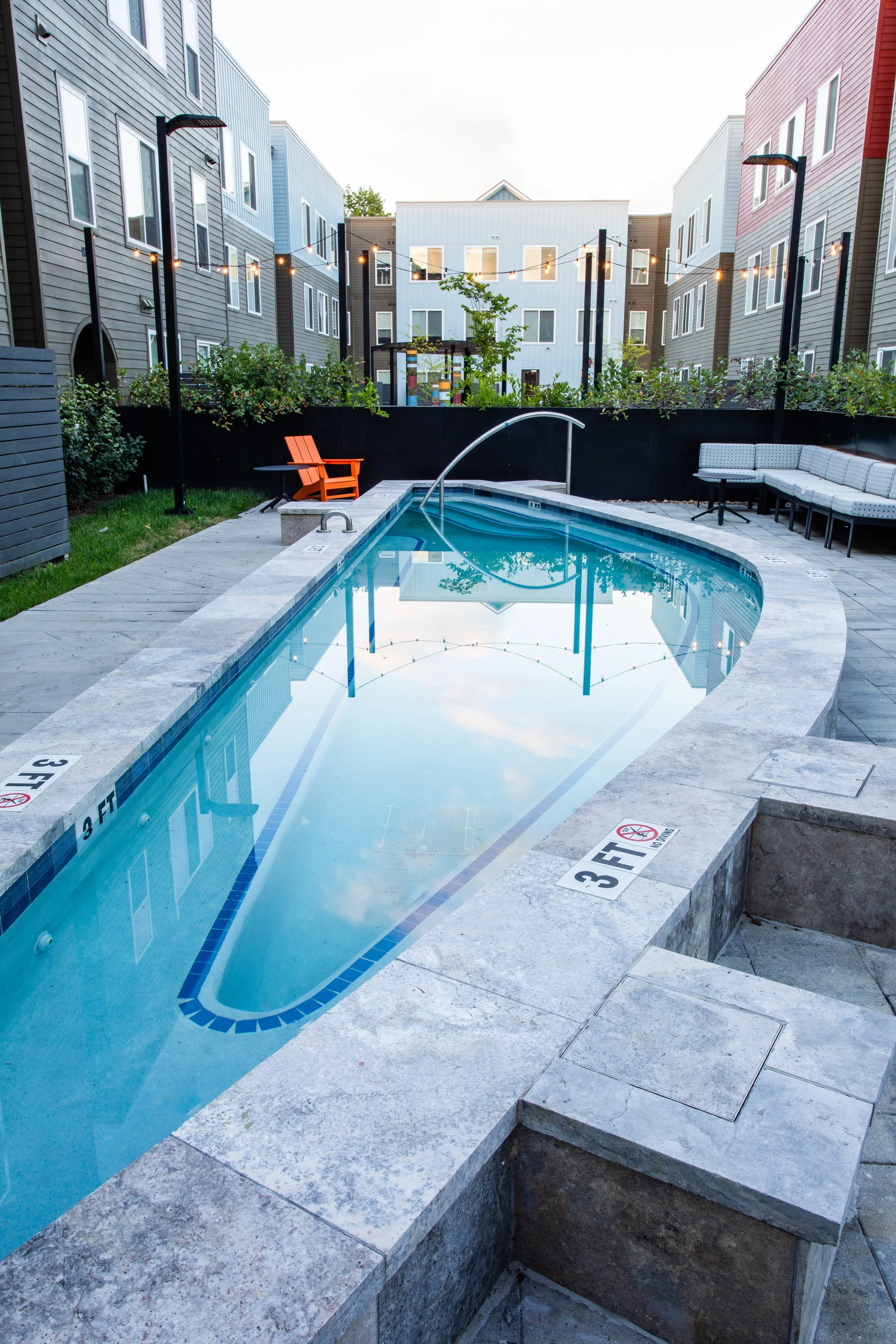 A small, outdoor swimming pool in an apartment complex courtyard, surrounded by modern buildings, with lounge seating and string lights overhead.