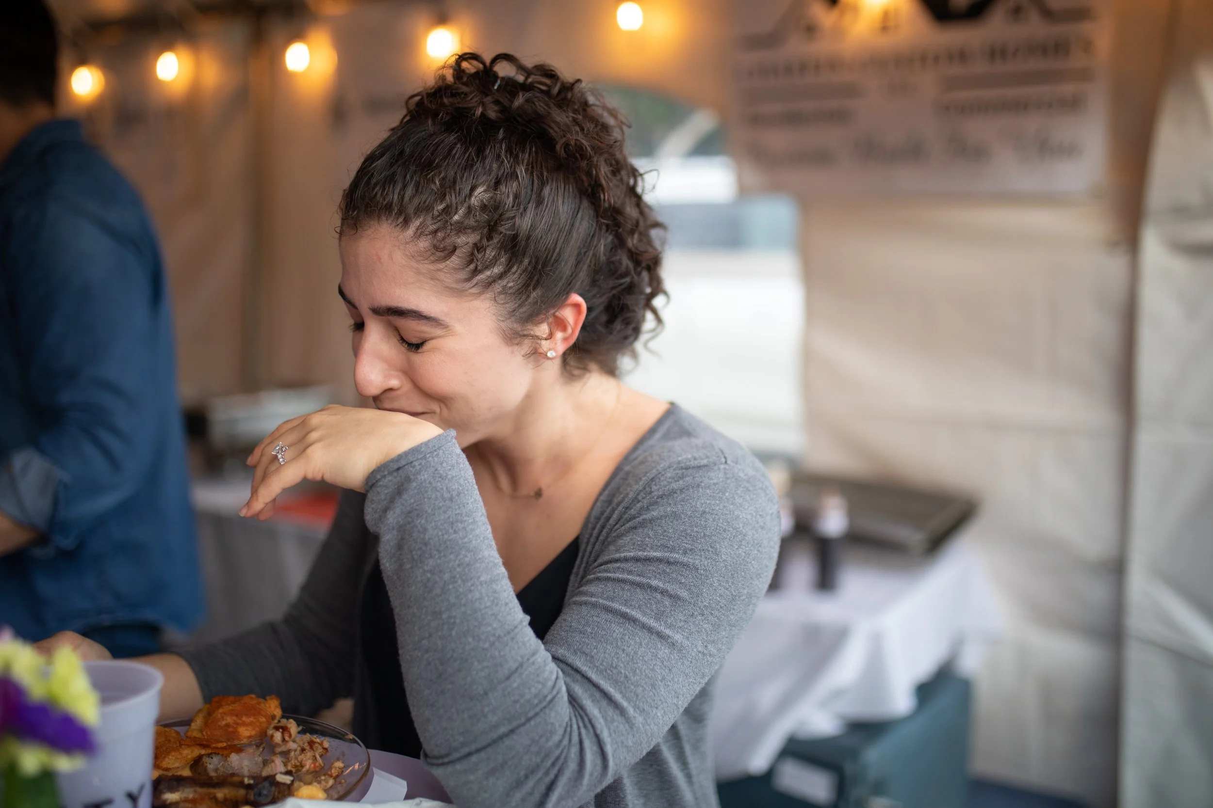 A young woman with curly hair smiling with her eyes closed, sitting at a table, appearing to be emotional or happy.