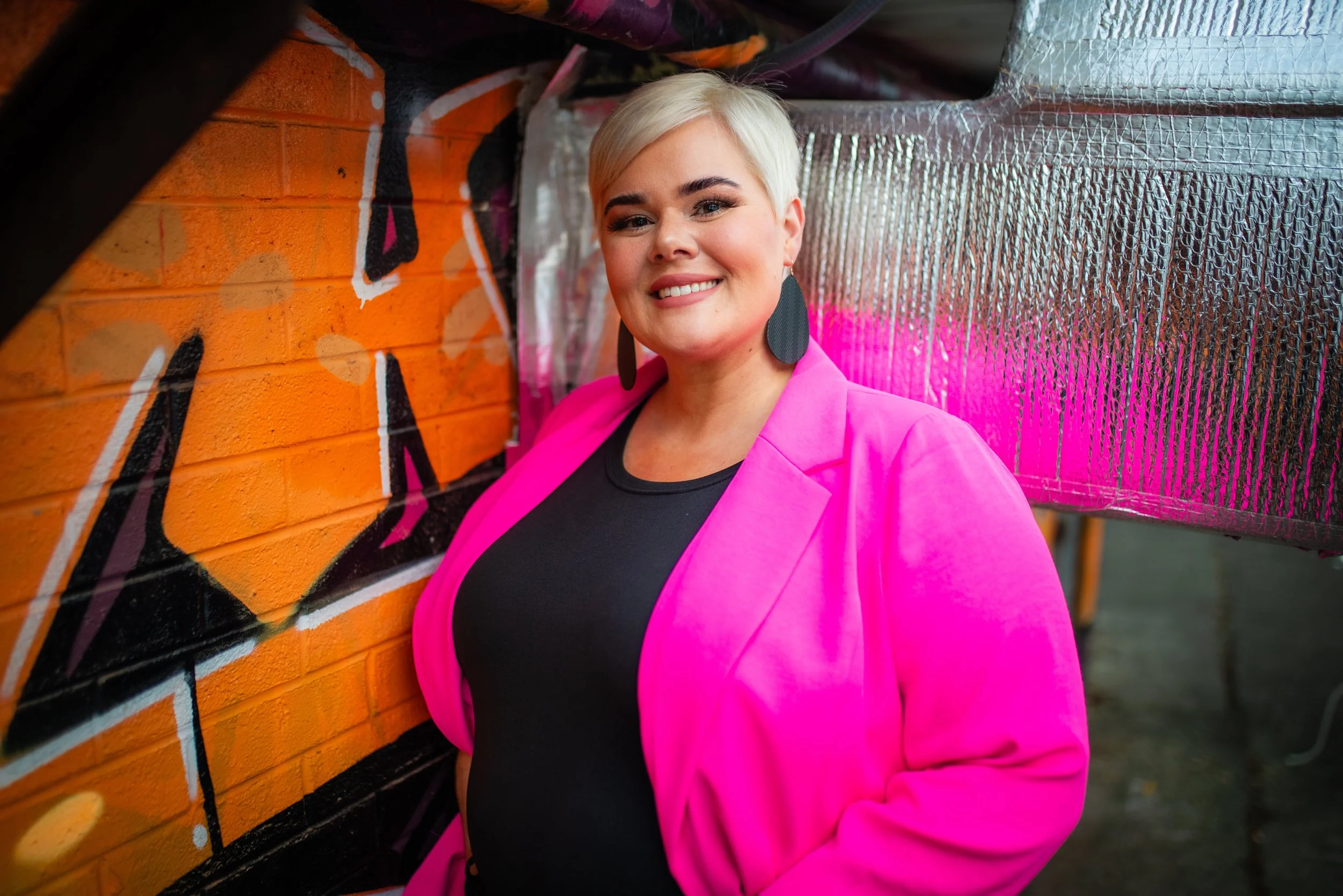 A woman with short blonde hair, wearing a pink blazer and large black earrings, smiling and standing next to a graffiti-covered brick wall with colorful artwork.