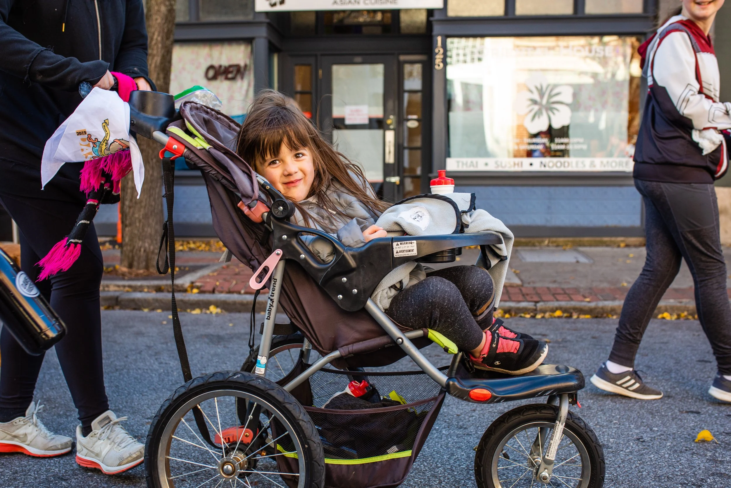 A young girl with long brown hair sitting in a stroller being pushed by an adult on a city street, with a restaurant in the background and other people walking by.