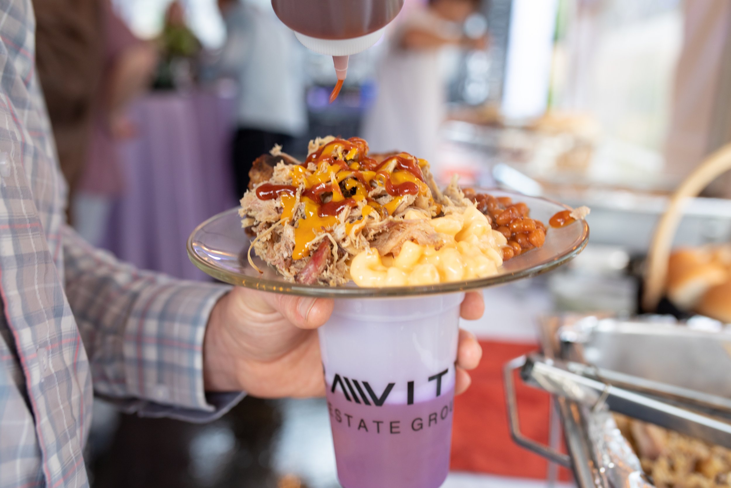 A person holding a clear glass plate with baked beans, pulled pork with shredded meat, and macaroni and cheese, with barbecue sauce and mustard drizzled on top, all served on a white cup labeled 'VIT'.