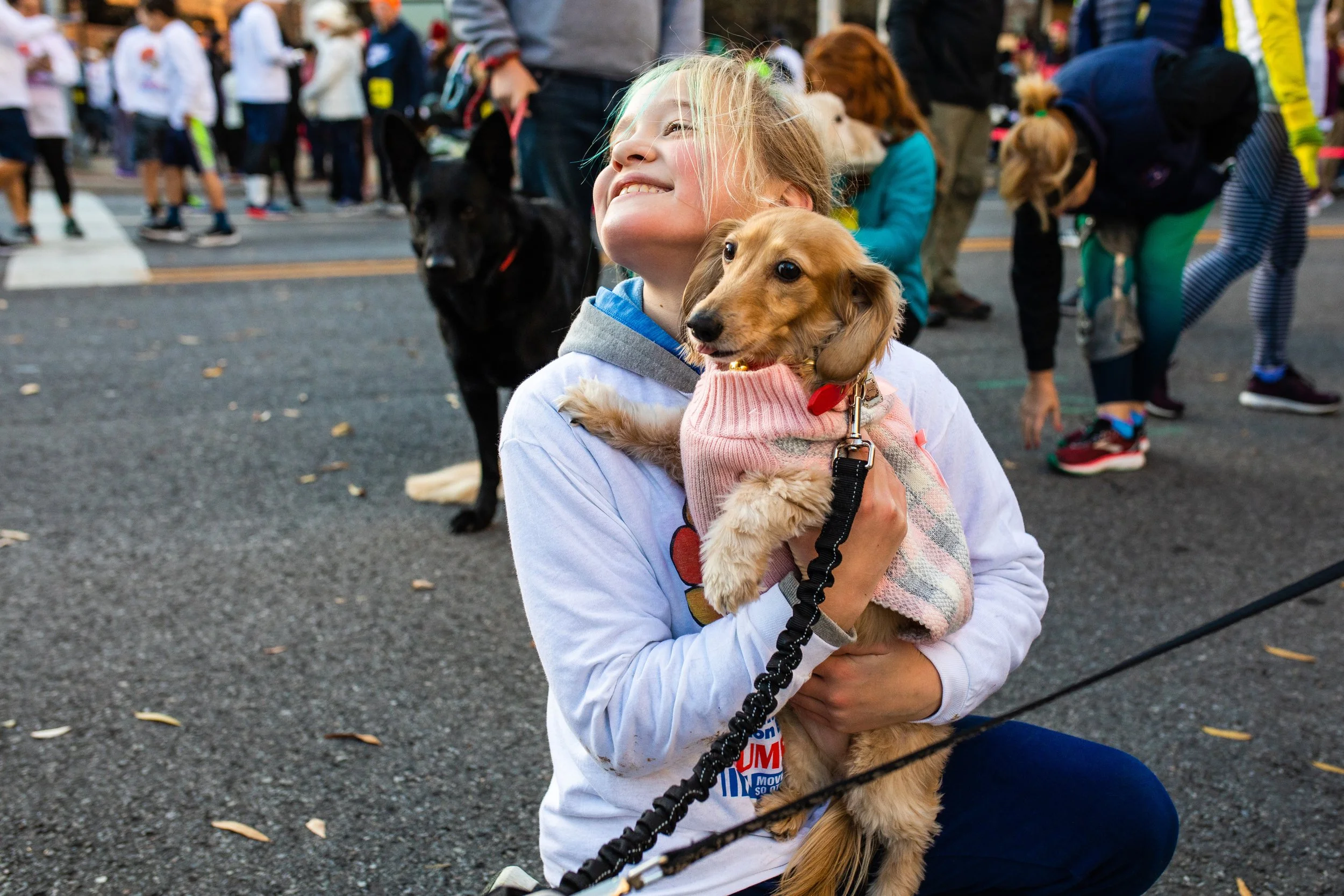 Young girl smiling and hugging a small dog at an outdoor event or walk, with a black dog and other people in the background.