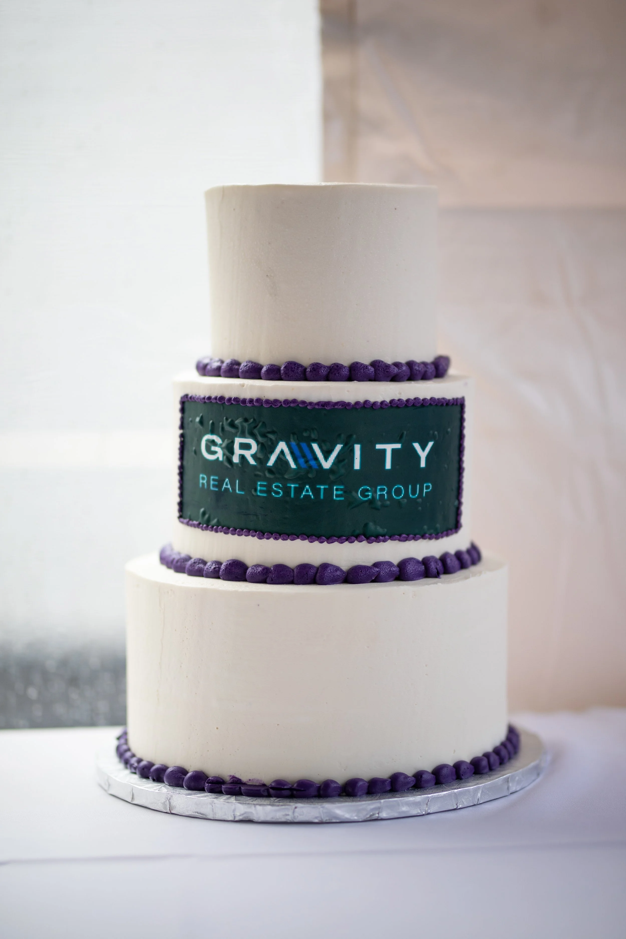 Three-tiered cake with purple piping and a logo that reads 'Gravity Real Estate Group' on the middle tier.