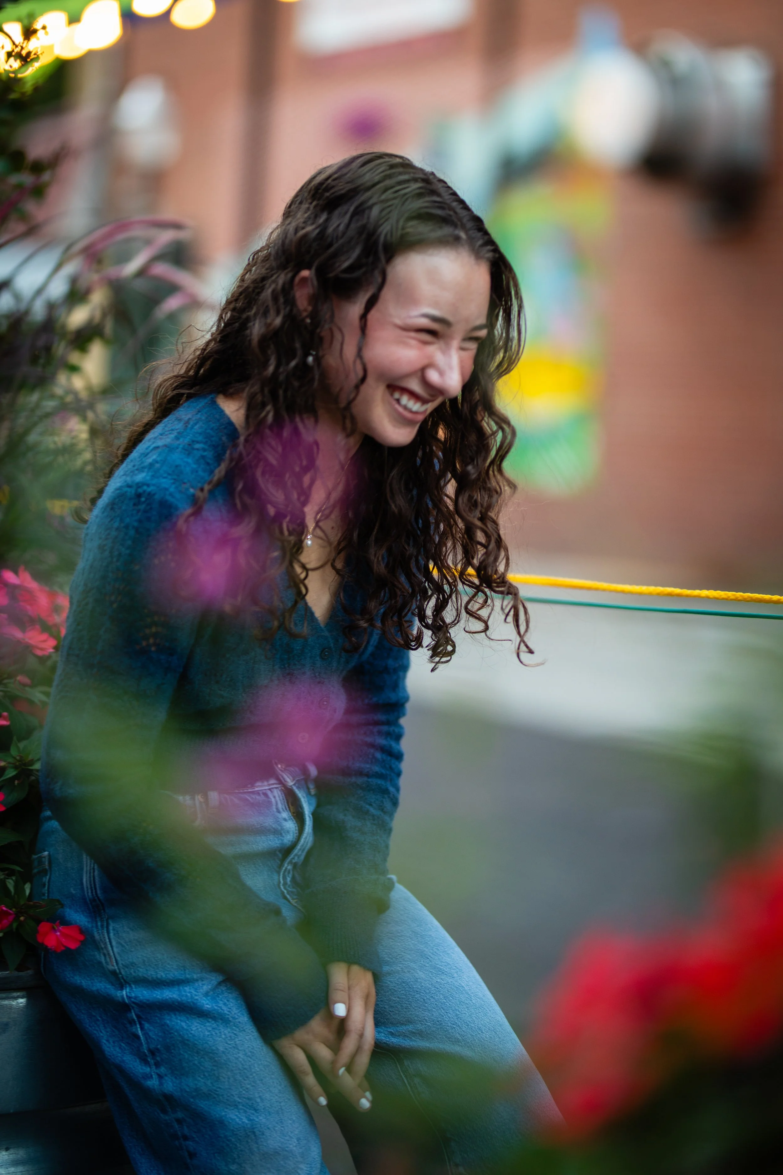 A woman with long curly hair, smiling and sitting outdoors near flowers with a blurred colorful background.